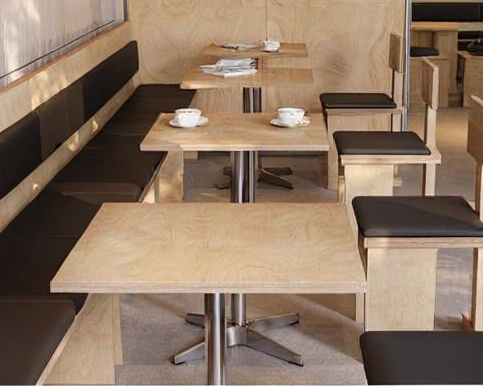 Empty cafe seating area with light wood tables, black cushioned bench seating, and black cushioned stools, with some cups and napkins on the tables.