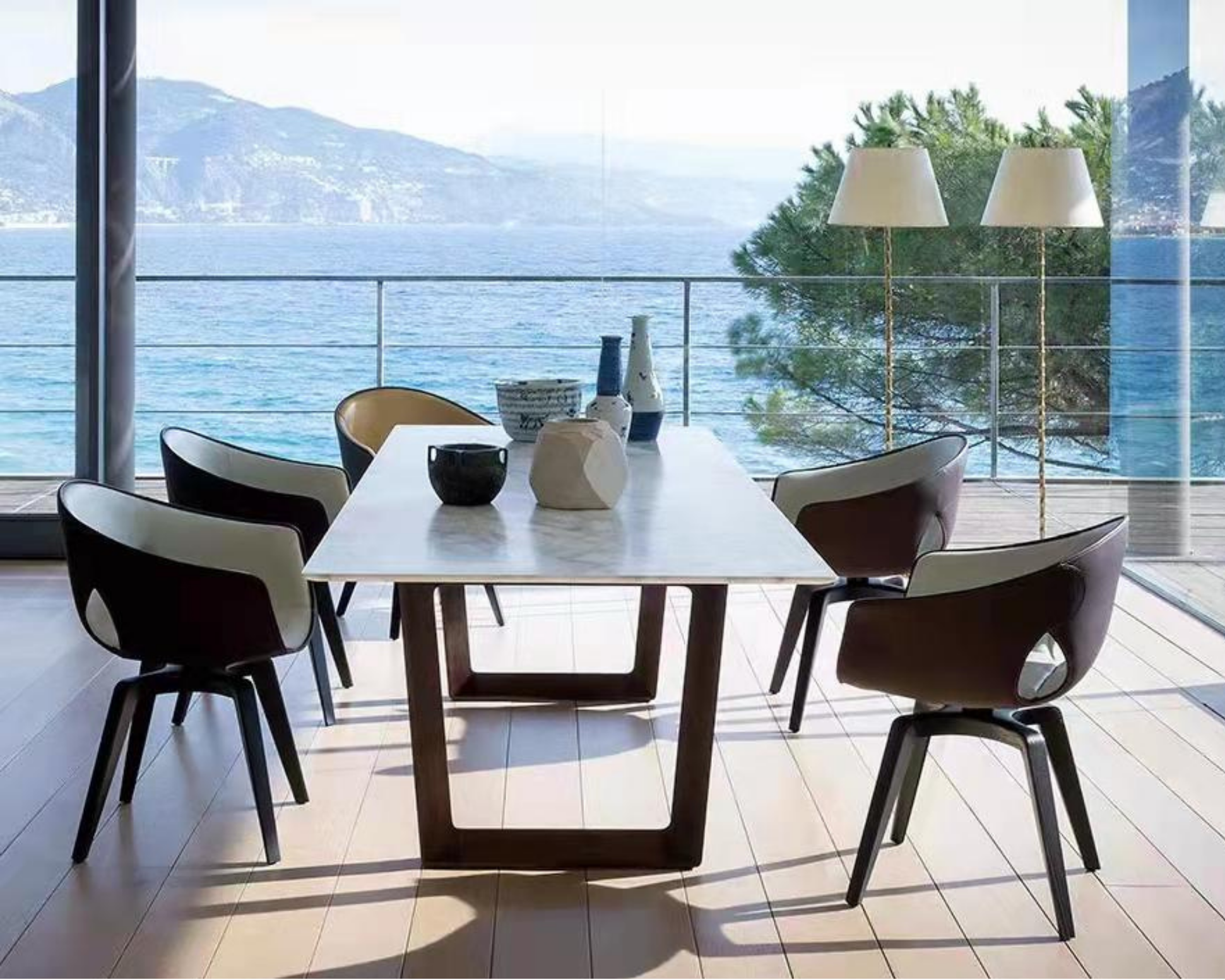 Dining area with a white marble table surrounded by chairs, overlooking a water body with mountains and trees visible through large glass windows, and decorated with vases and lamps.