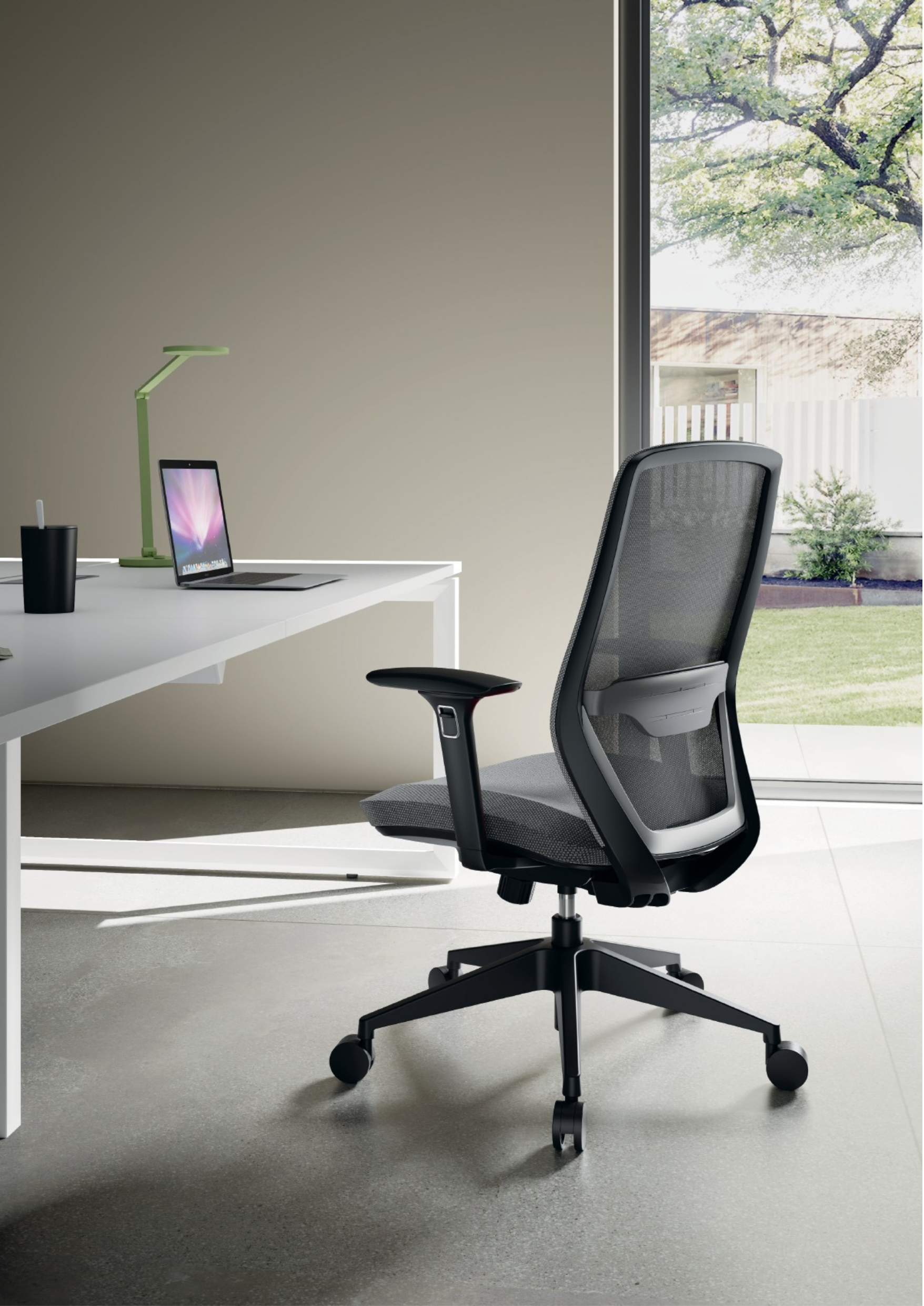 Modern office workstation with a black ergonomic chair, white desk, green desk lamp, black cup with a white pen, and a laptop. Large window shows trees outside.