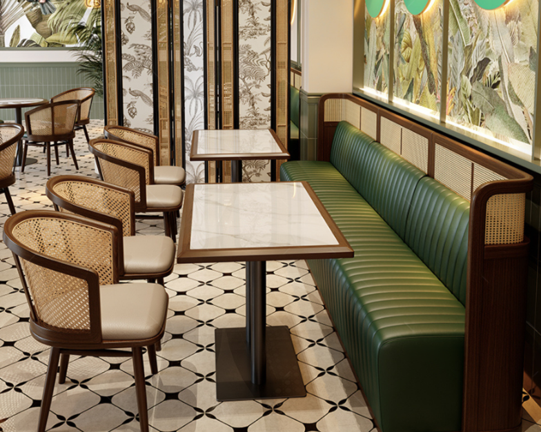Interior of a café with green bench seating, wooden tables with marble tops, and chairs with rattan backs and beige cushions, tiled floor, and tropical wall decorations.