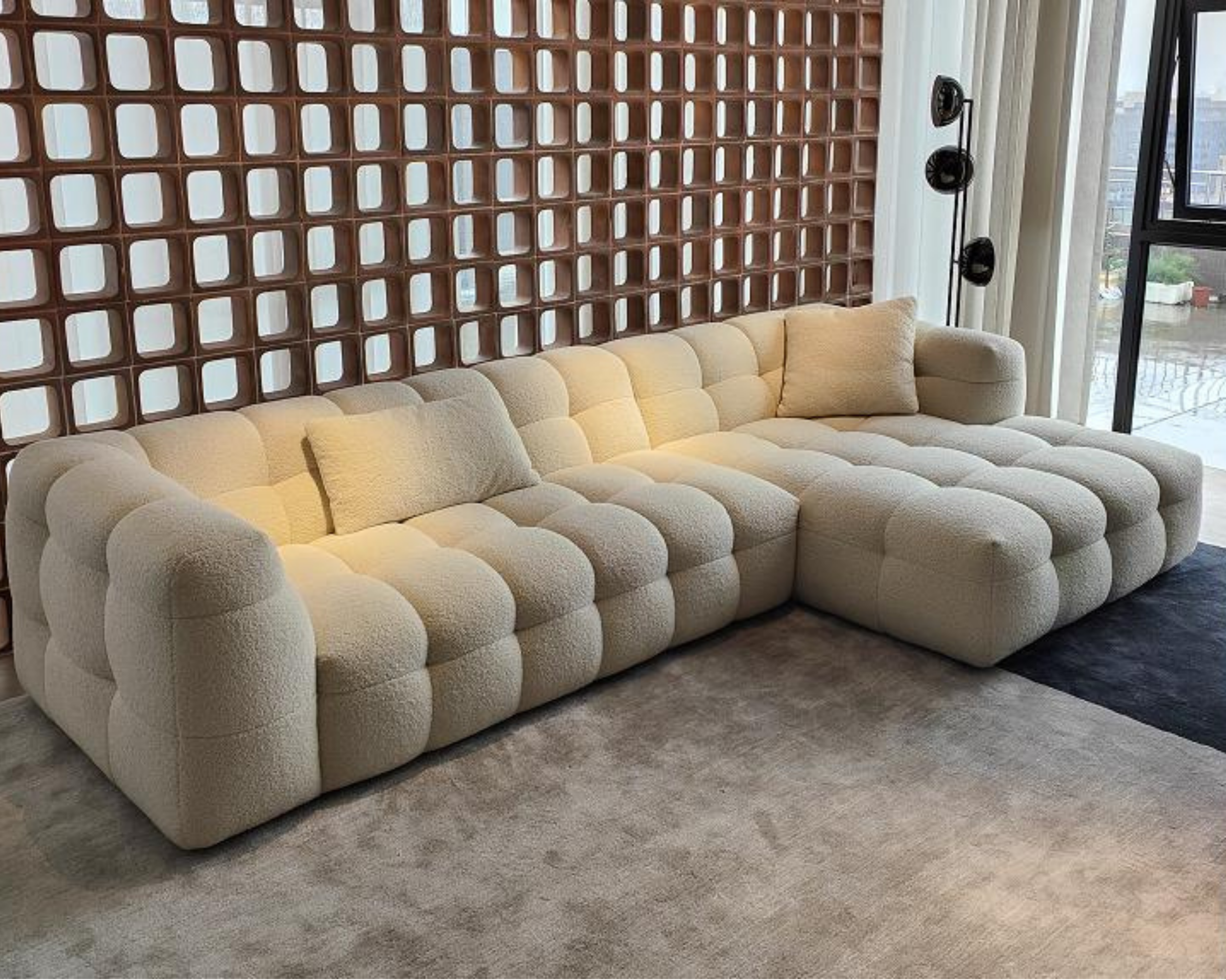A beige, tufted sectional sofa with rounded edges on a light gray carpet in a living room. Behind the sofa is a decorative brown partition wall with square openings, and to the right, white curtains partially cover a large window with a city view.