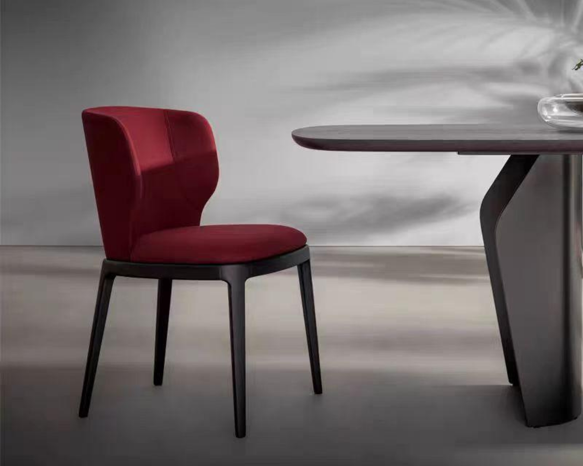 A red modern chair with a rounded backrest and black legs positioned next to a dark wooden table with a curved edge in a minimalistic setting with a light abstract background.