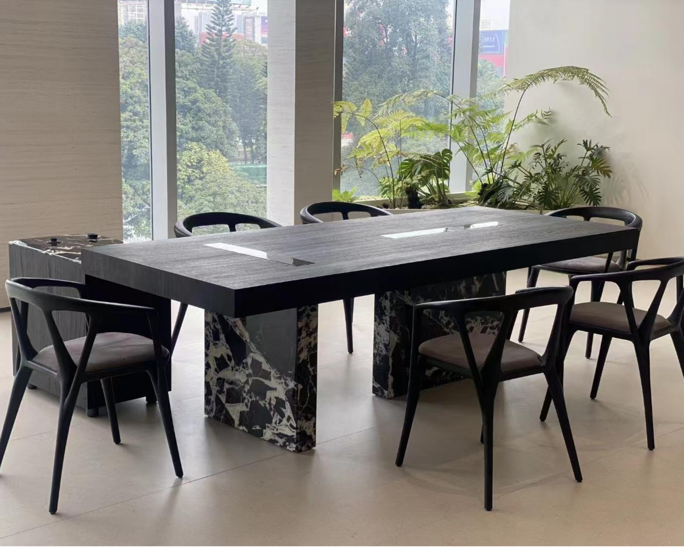 Modern meeting room with large windows overlooking city trees. Centered black table with marble legs, surrounded by six black chairs. Green leafy plants on window sill. Light-colored floor and plain walls.