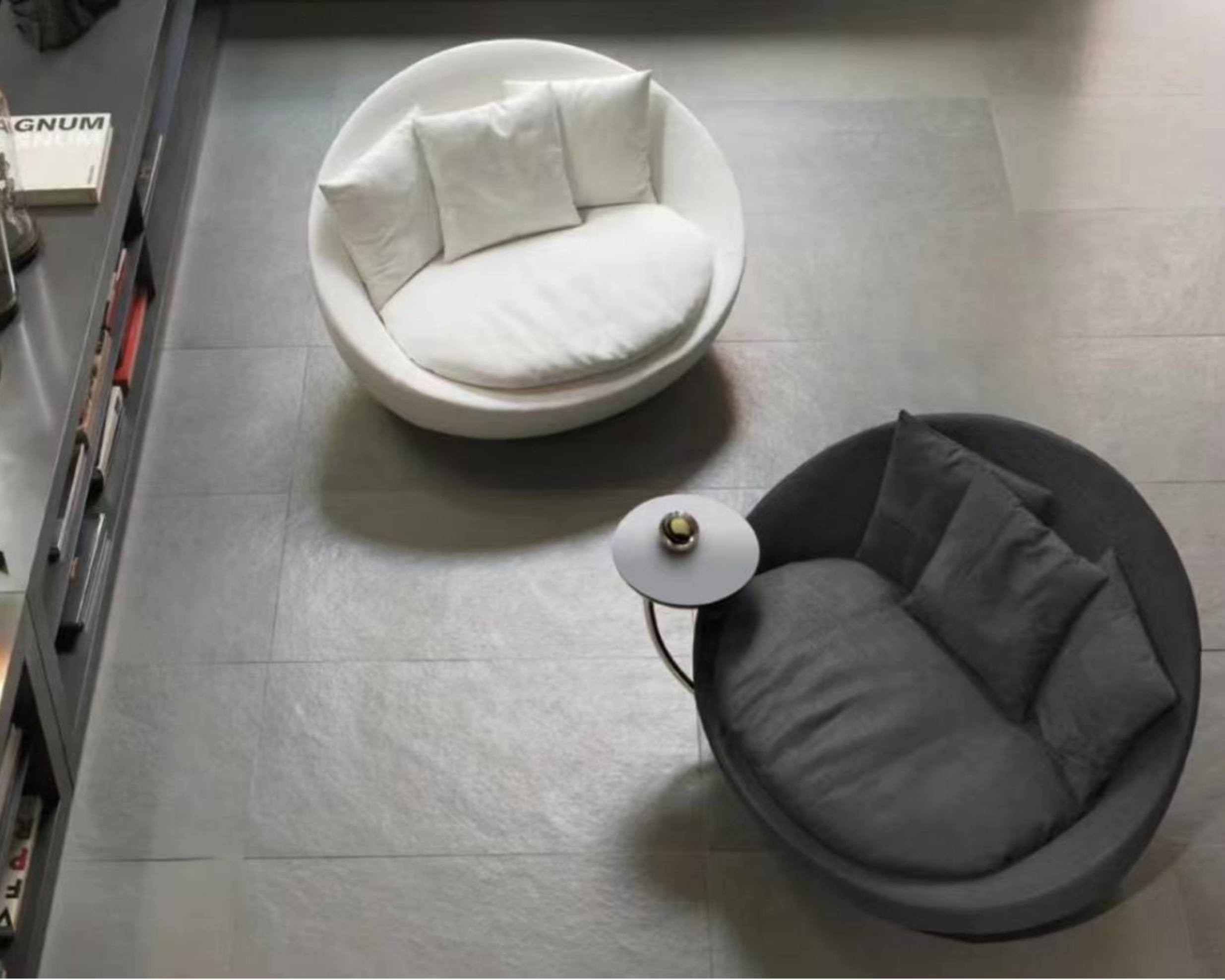 A white, round chair with cushion and pillows, positioned on a gray floor near a bookshelf and a table with a small lamp on it.