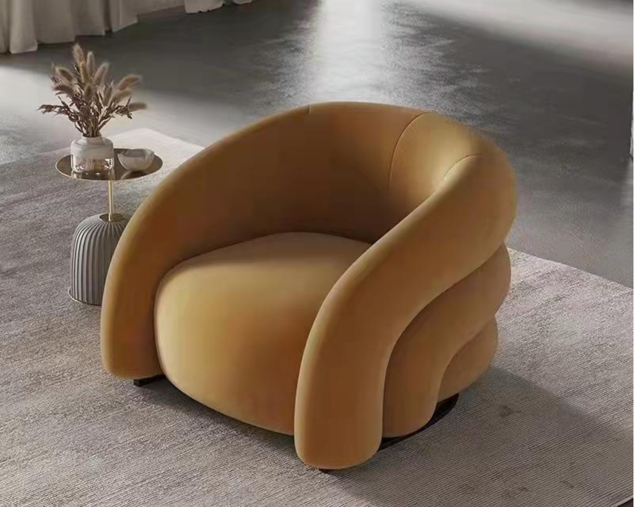A modern, curved, mustard-colored armchair on a neutral-toned carpet near a side table with a white vase holding dried pampas grass and a small bowl. The background features a polished concrete floor and curtains.