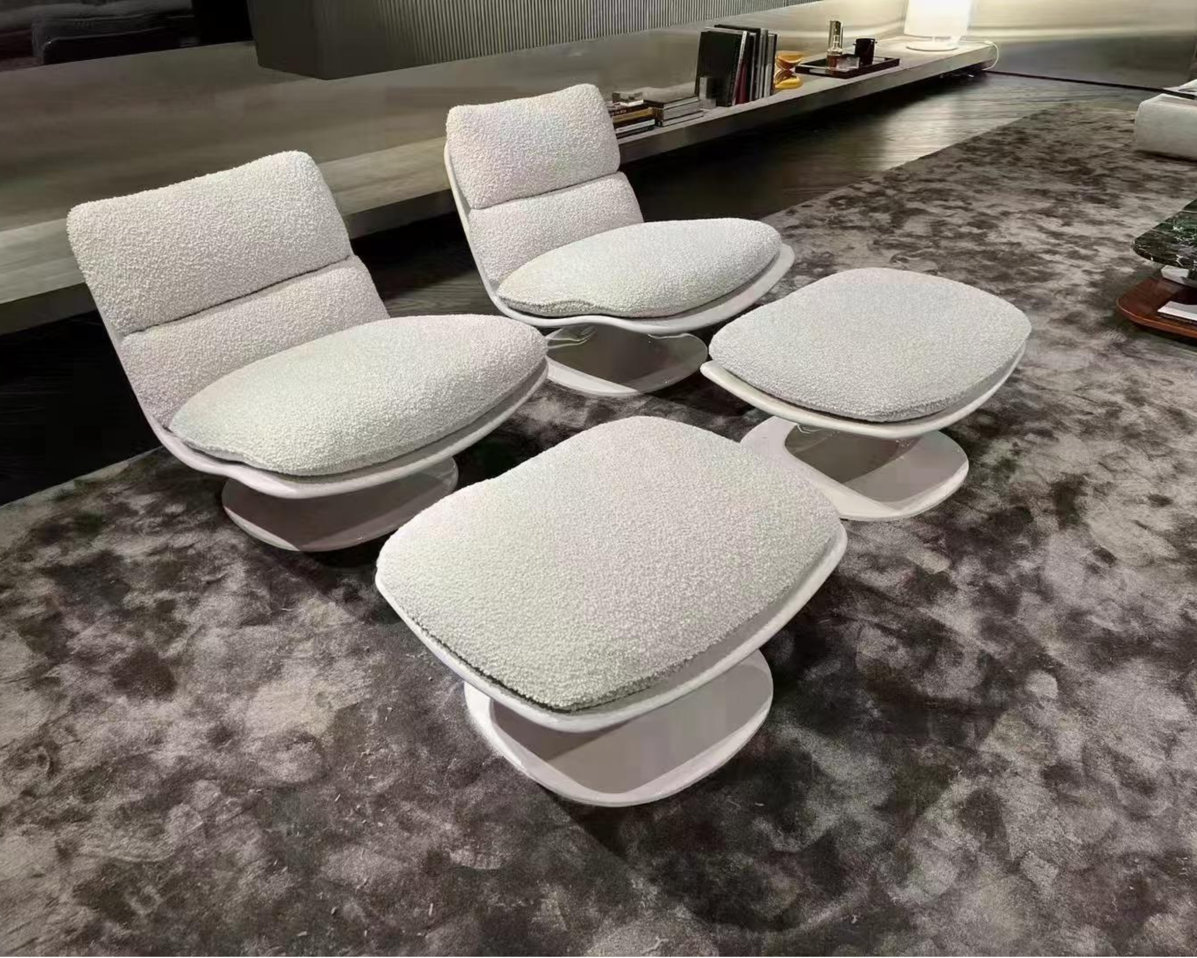 A modern living room with three white textured lounge chairs and a round ottoman set on a patterned rug, with a long console shelf in the background holding books and decorative items.