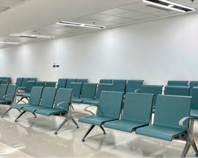 Empty airport waiting area with teal chairs arranged in rows, white walls, and fluorescent ceiling lights.