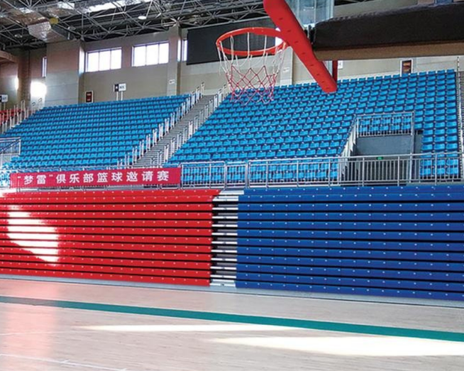 Empty indoor basketball court with blue bleachers, red and blue stairs, and a basketball hoop with a red rim.