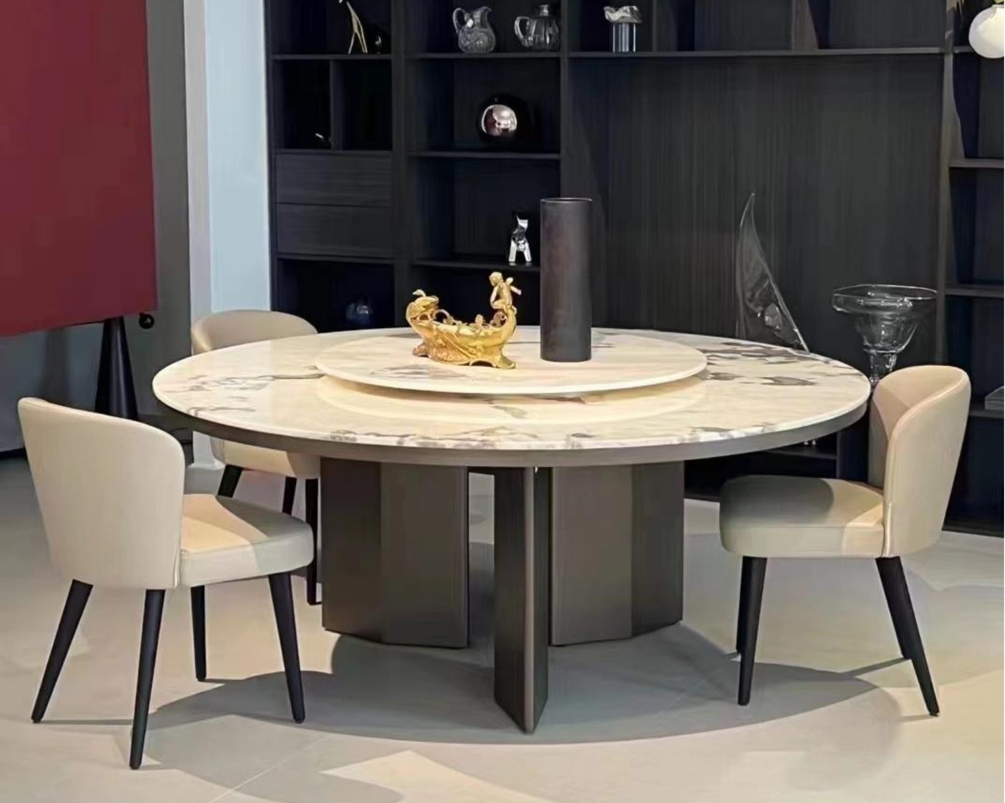 Round dining table with a marble top, surrounded by six white chairs with black legs. Decor items on the table include a large black cylindrical vase, a small carved gold sculpture, and a clear glass bowl. In the background, a black shelving unit displays various decorative objects, and part of a red wall and a white wall are visible.