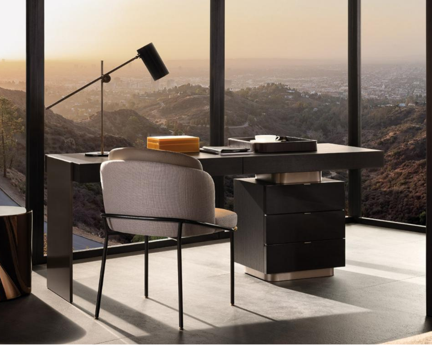 Modern office desk with a beige chair, a black desk lamp, and cityscape view through large windows at sunset.