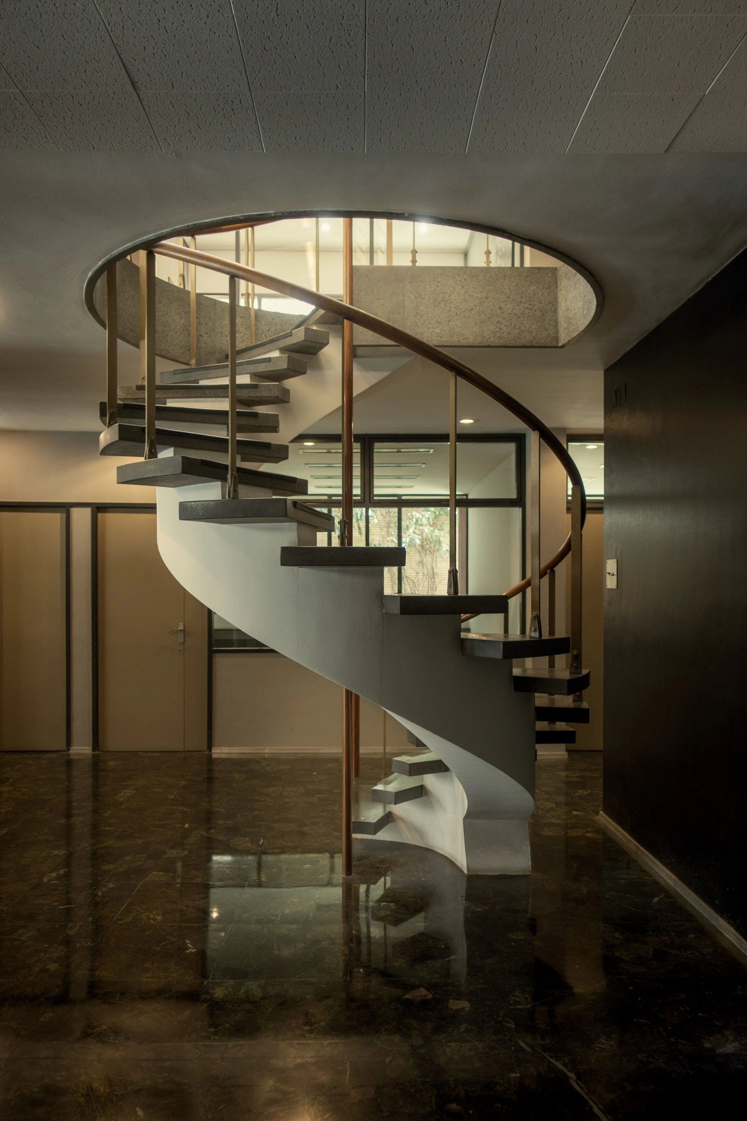 Escalier en colimaçon en béton dans un bâtiment moderne, avec un plafond à tuiles, murs en pierre, sols en marbre foncé, fenêtres en verre, et un mur noir sur le côté