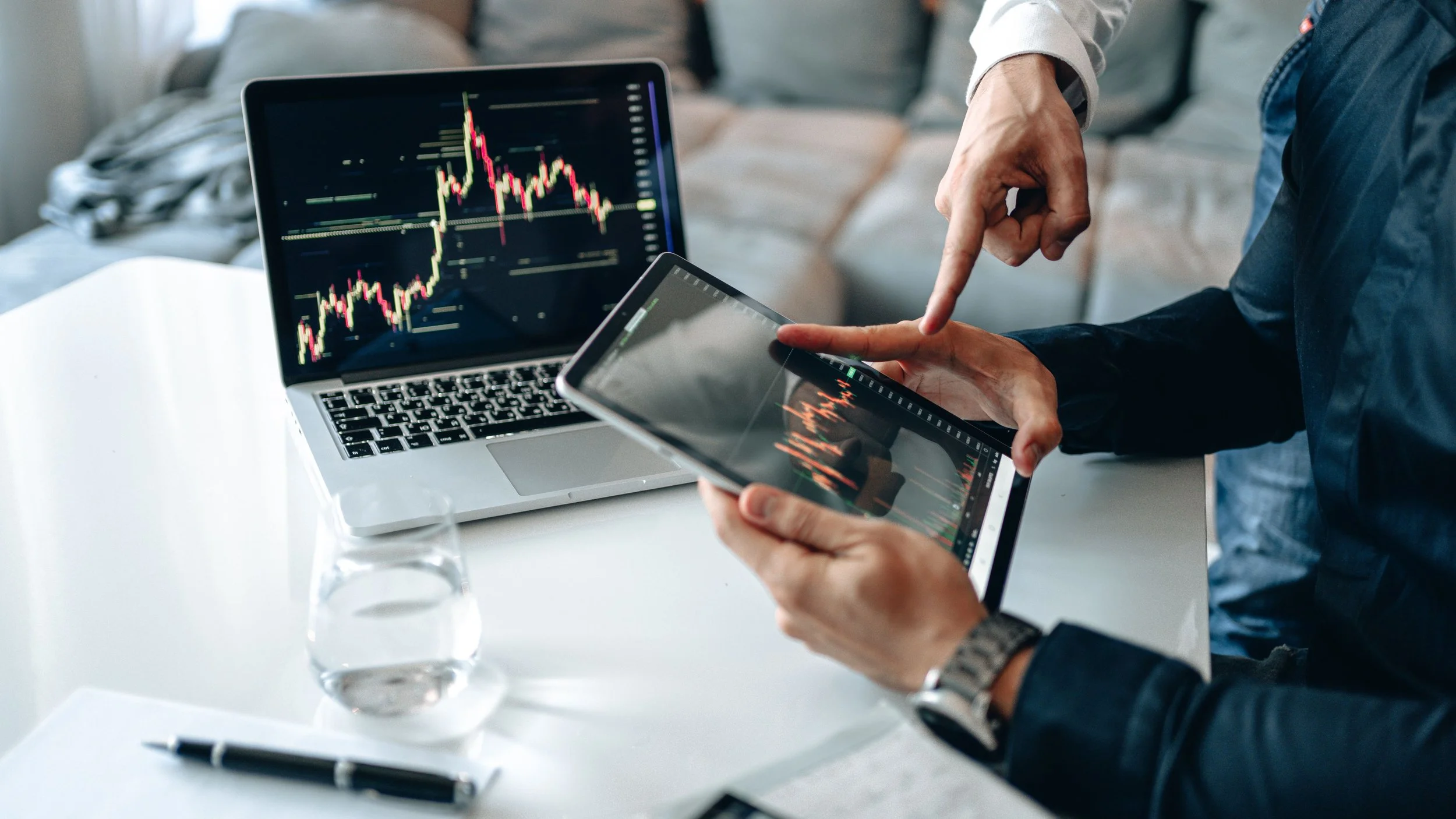 Deux personnes analysent des graphiques de trading sur un ordinateur portable, une tablette et une feuille sur une table, dans un environnement professionnel.