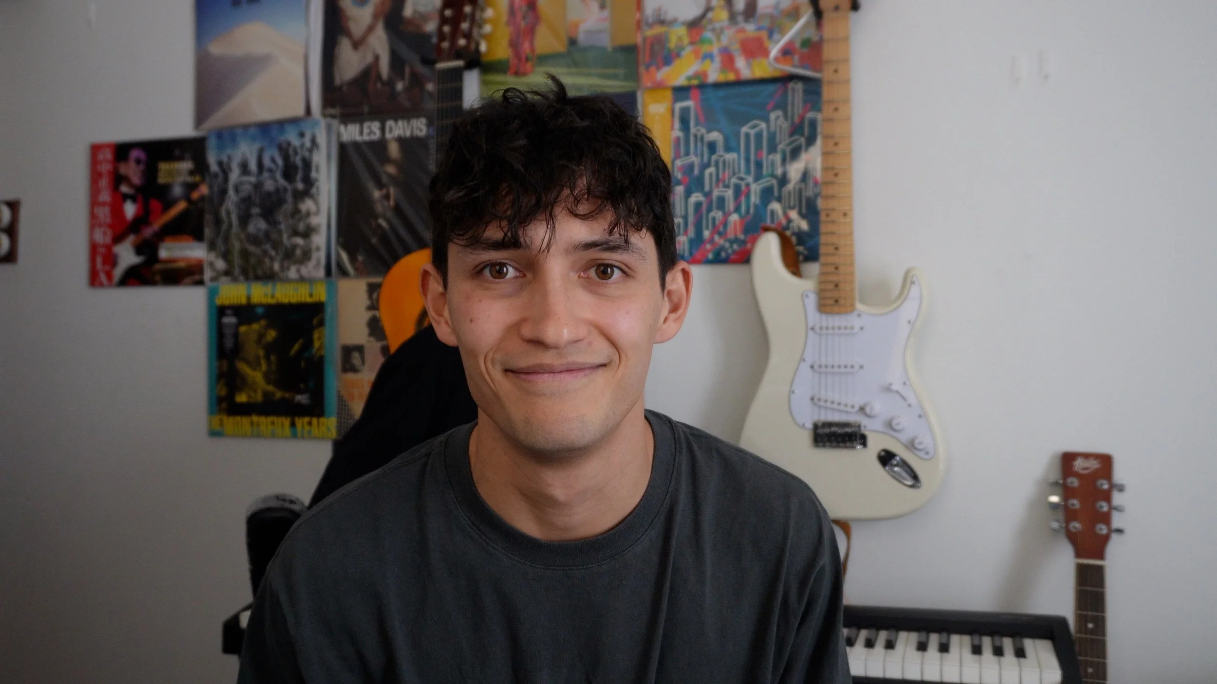Young man with dark curly hair smiling at the camera in a room with musical posters, a white electric guitar, and a keyboard in the background.
