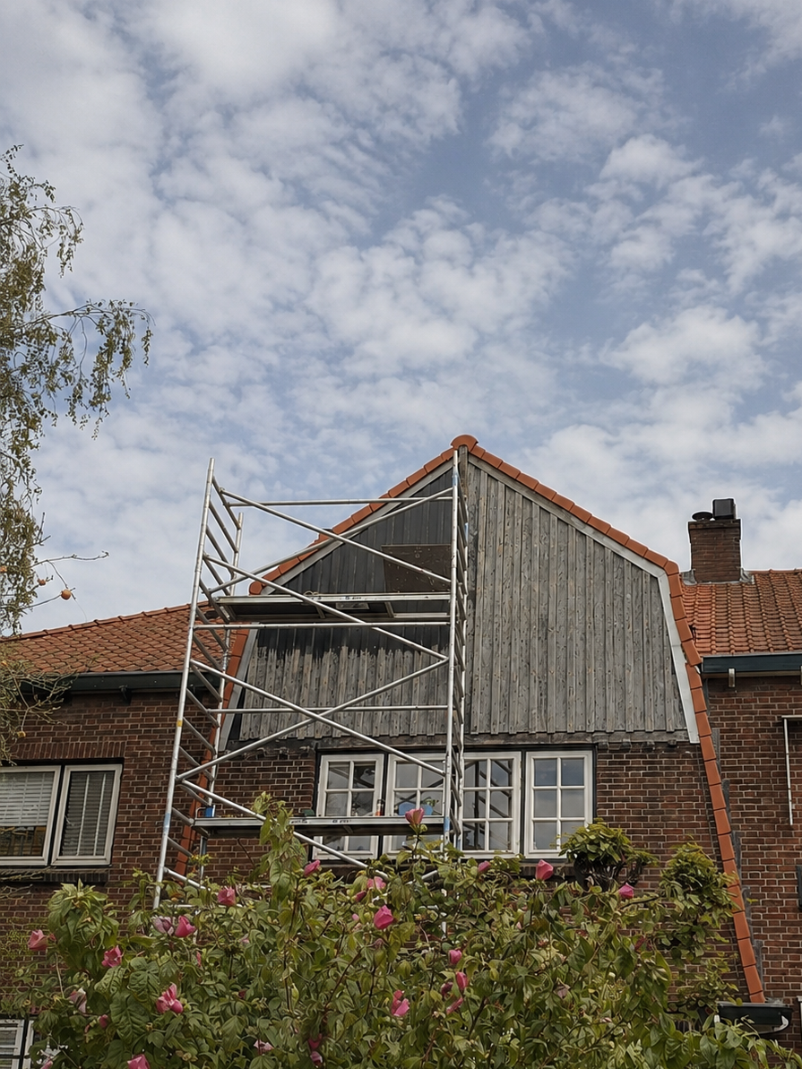 Een huis met een steile dakrand, een houten gevel, een schoorsteen en een bloemenstruik voor het huis. Op het dak staat een steiger die waarschijnlijk wordt gebruikt voor onderhoud of renovatie.