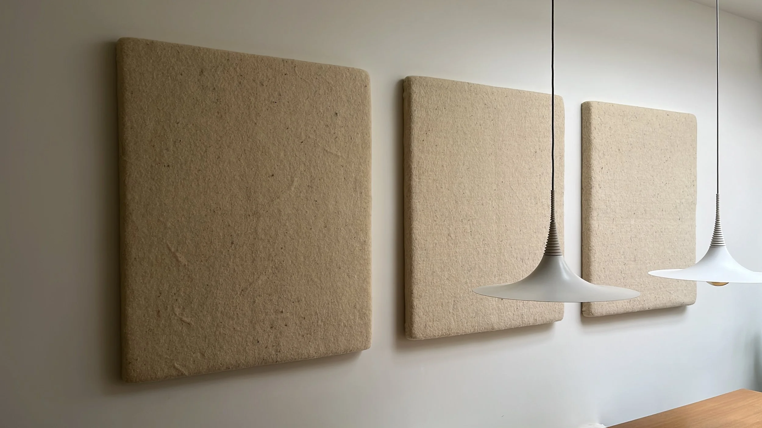 Drie beige geluiddempende panelen aan een witte muur met twee hanglampen eronder.