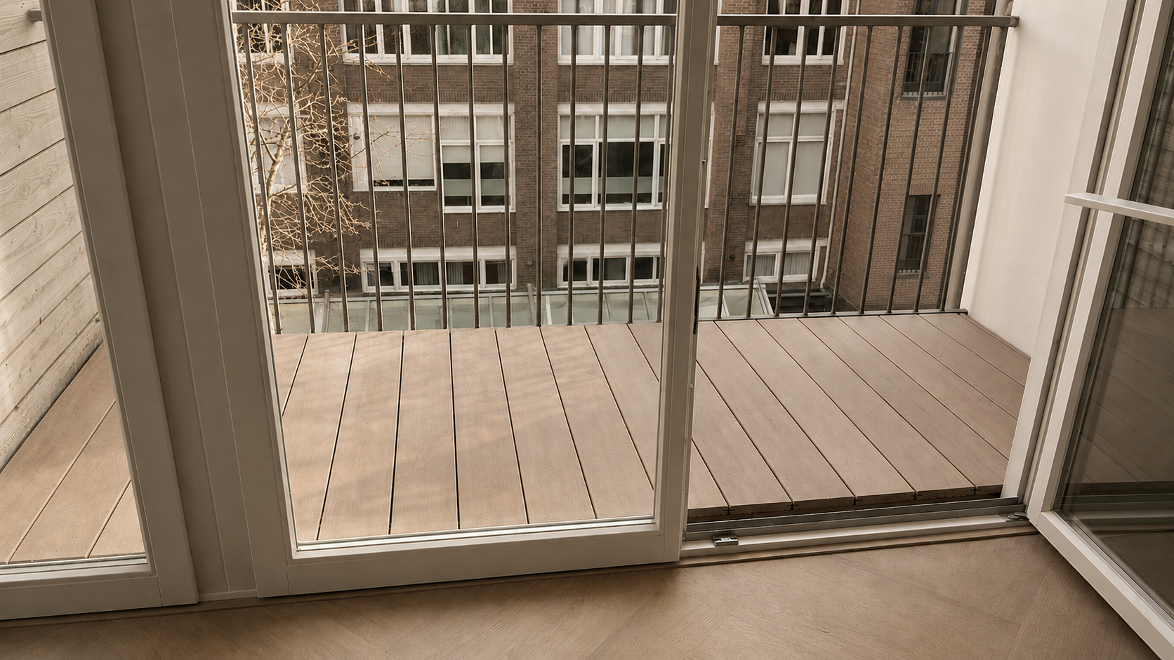 Een klein balkon met houten vloer en metalen railing, uitzicht op een gebouw met ramen en een bladloze boom.