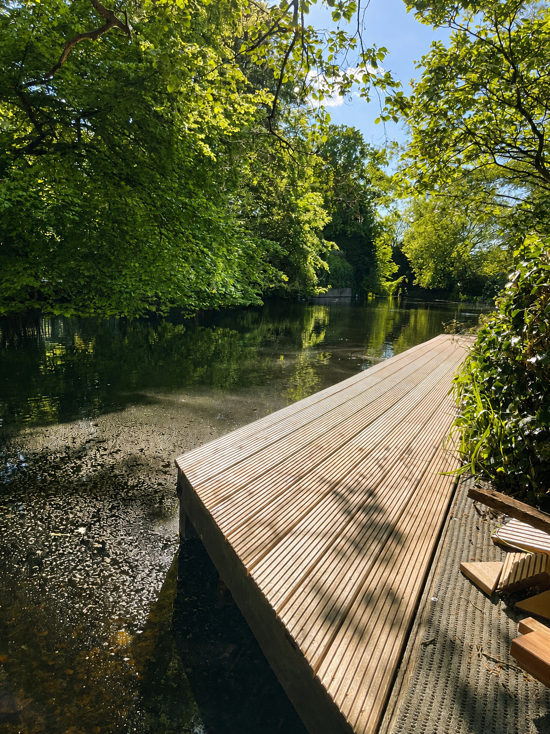 Een houten steiger langs een rustige, met water gevulde rivier omringd door groene bomen onder een heldere blauwe lucht.