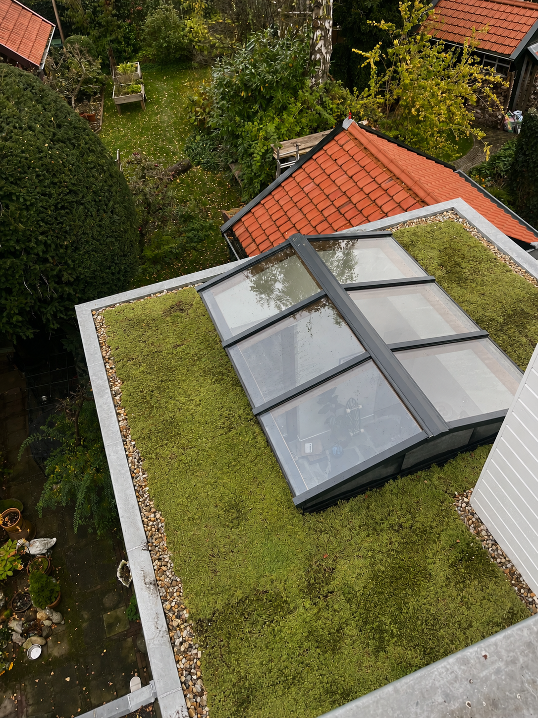 Bovenaanzicht van een groendak met mos en grindranden, met een glazen luifel en omliggende tuin met bomen en struiken.