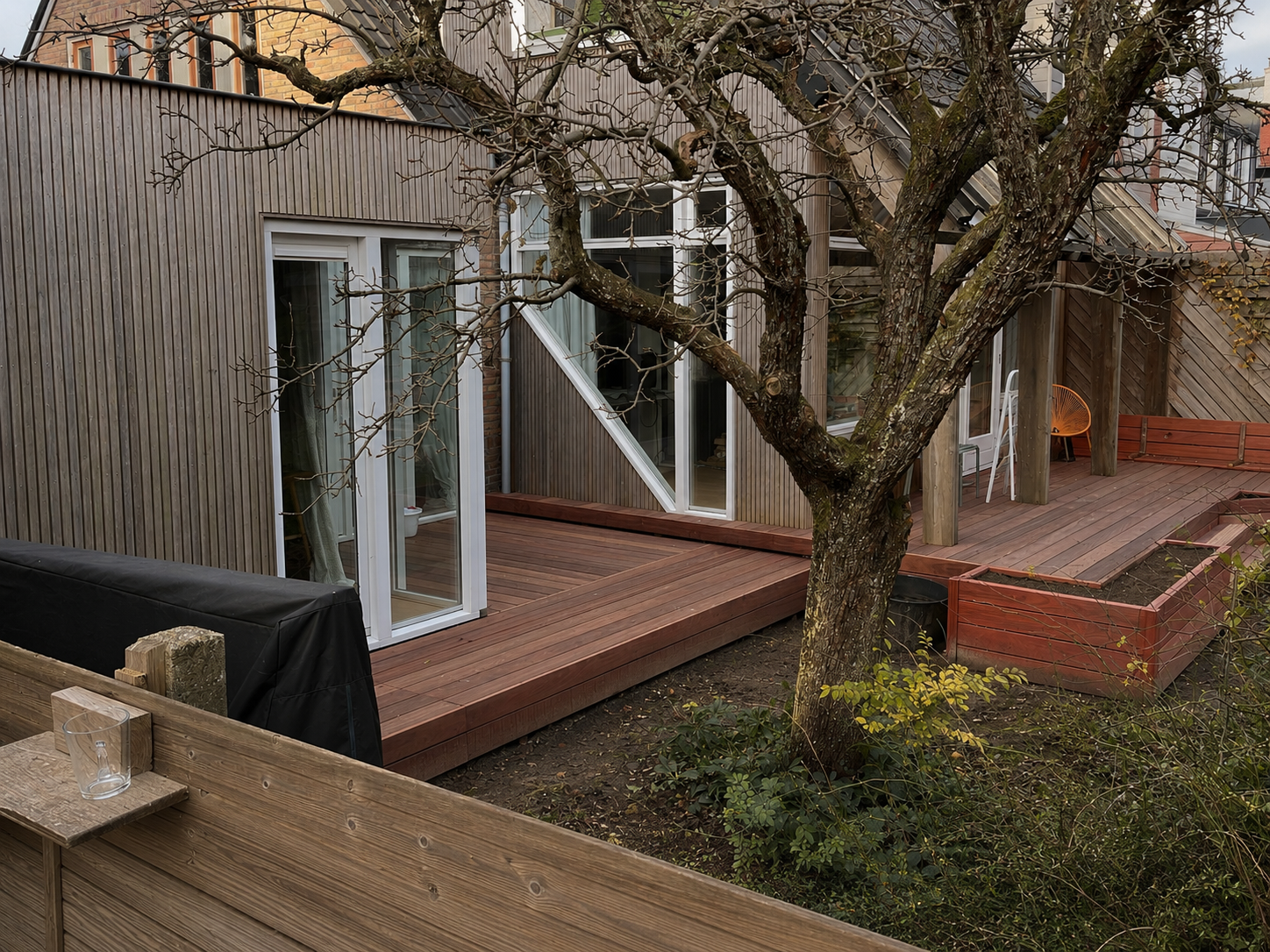 Een nieuwe houten terras aangebouwd aan een huis met grote glazen schuifdeuren, omringd door een tuin met een grote, bladloze boom en een houten afscheiding.
