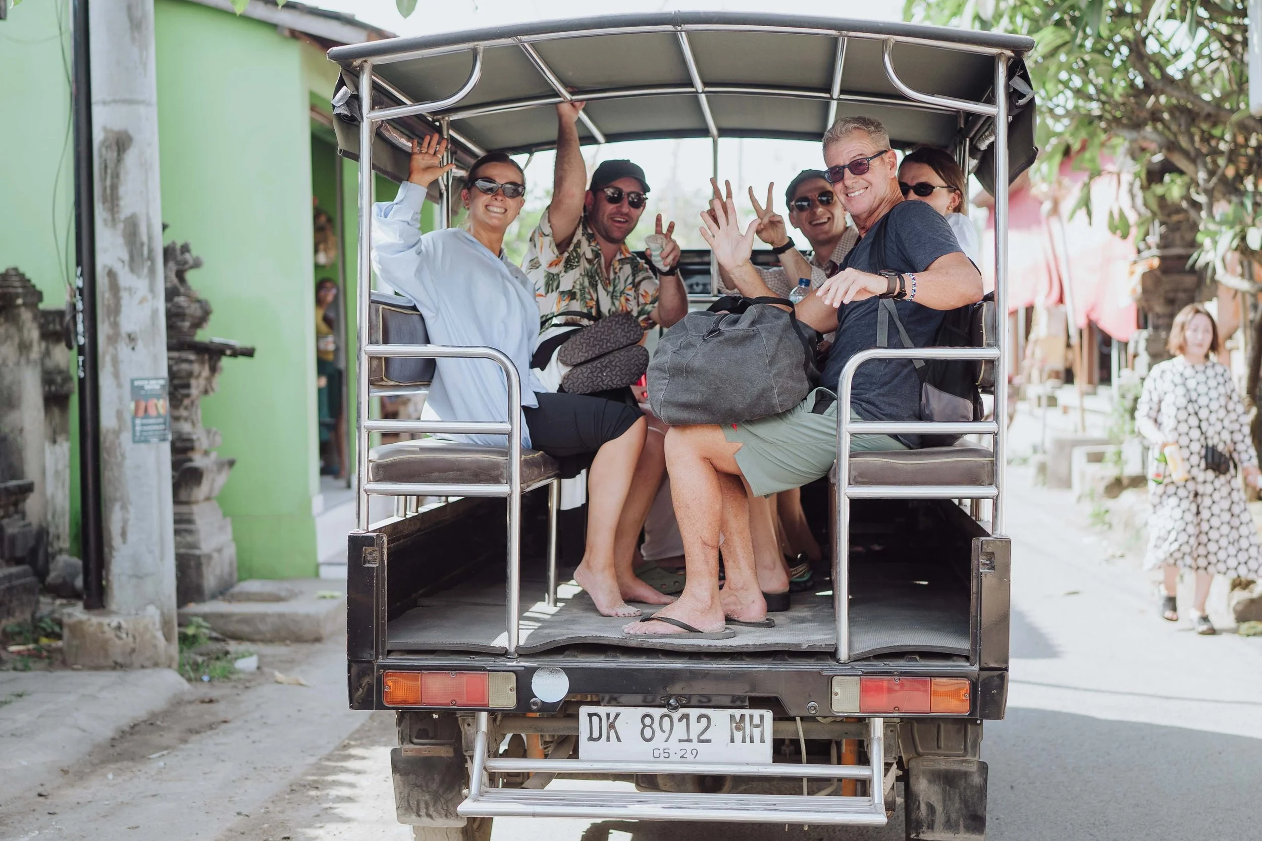 A group of six people smiling and waving while riding in a small open truck on a street in a tropical or warm climate area.