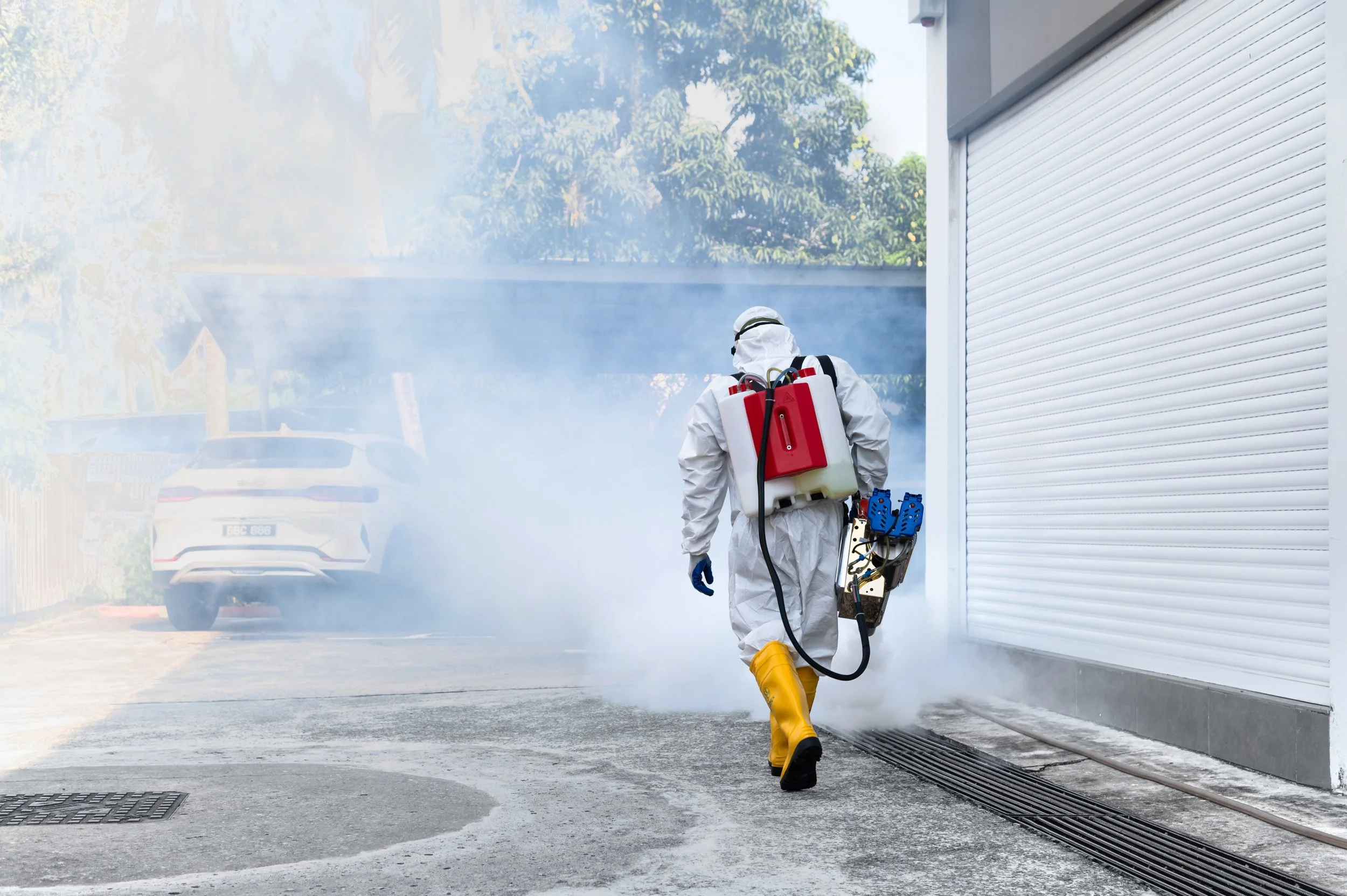 Person wearing protective suit and yellow boots spraying disinfectant near a white industrial building, surrounded by a haze of disinfectant spray.