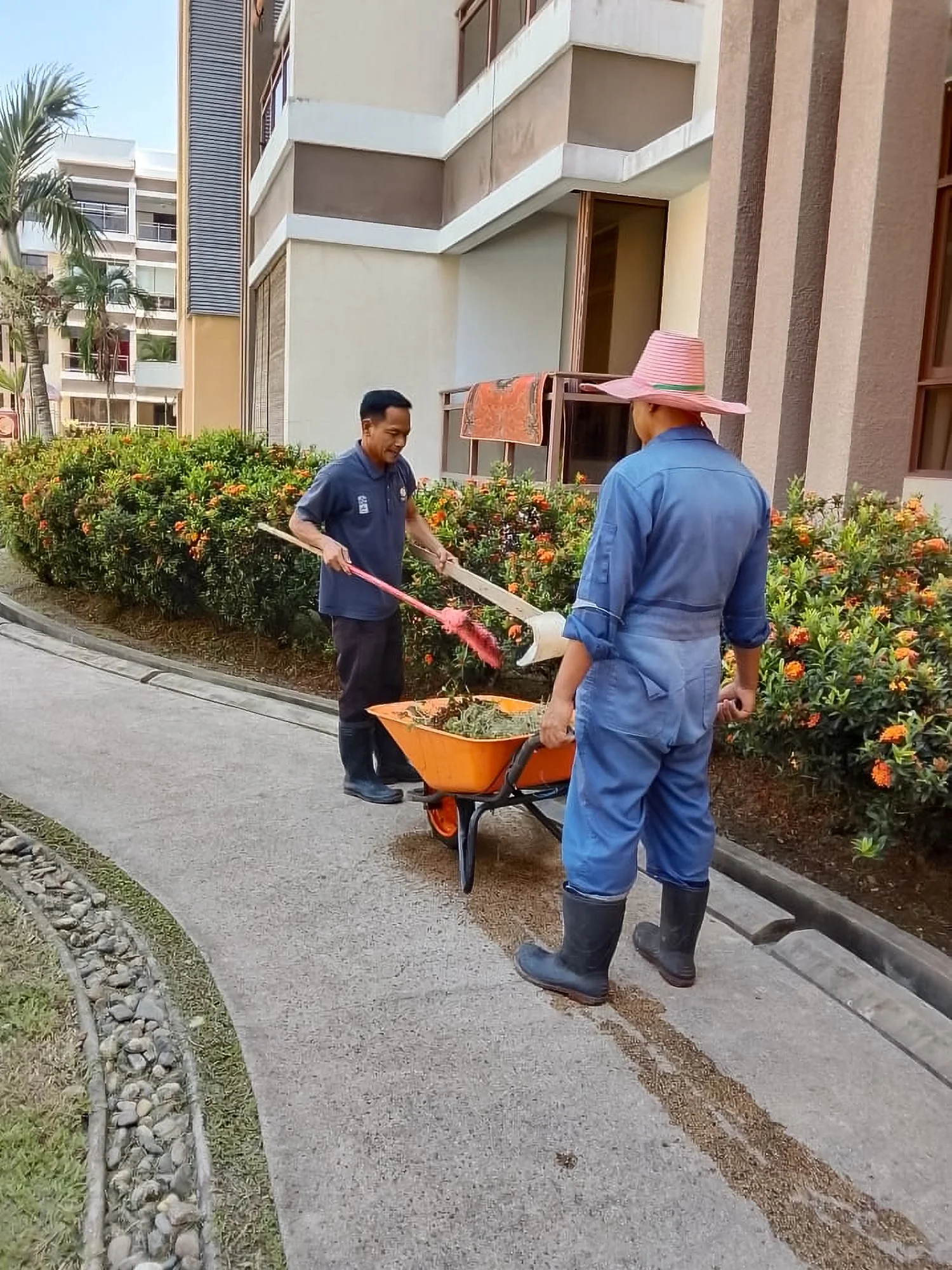 Ground maintenance services outside a Brunei apartment building using a wheelbarrow and rake for property upkeep.