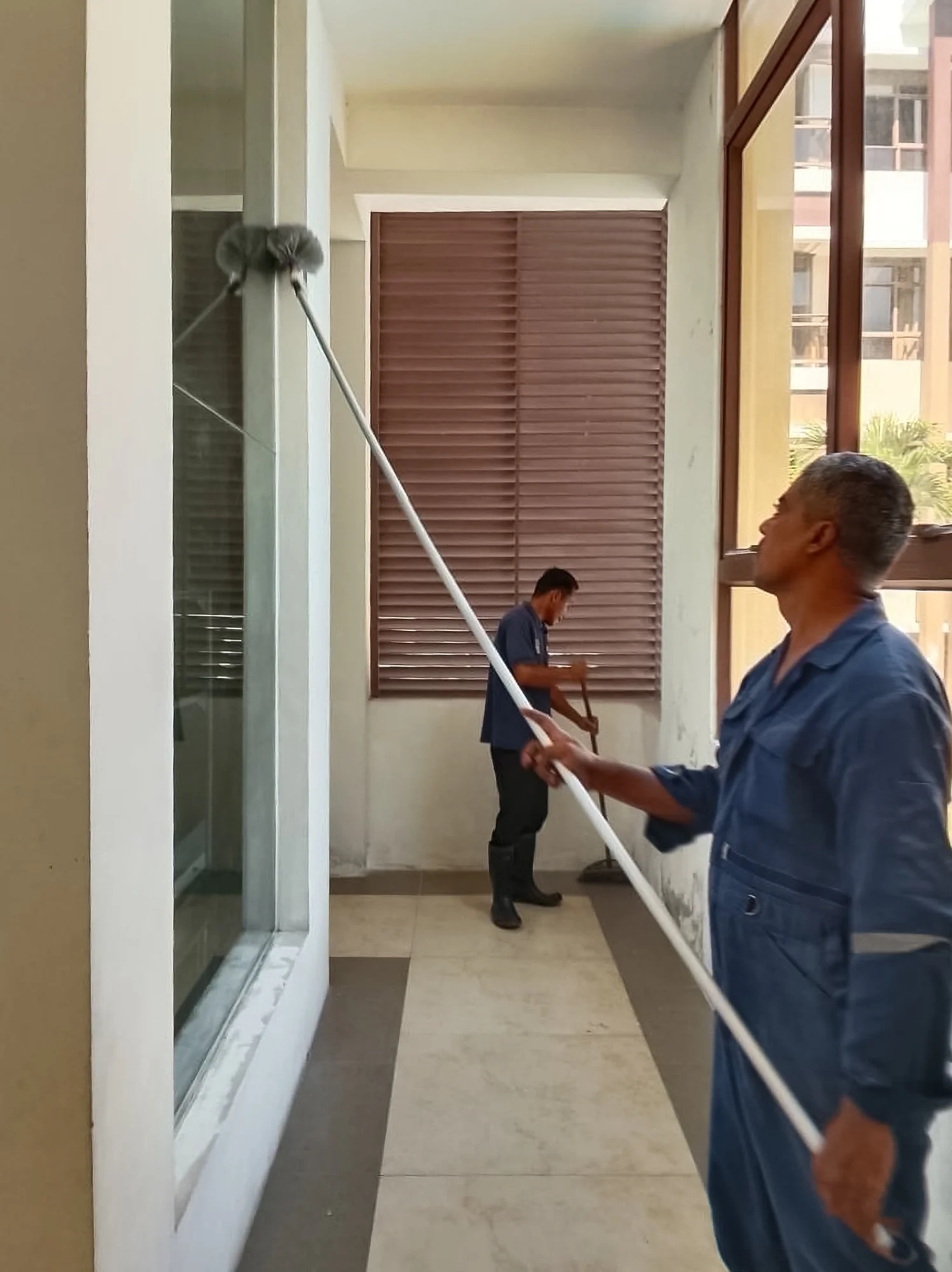 Two maintenance workers cleaning a building window with a long-handled cleaning tool inside a corridor near large windows and wooden shutters in Brunei.