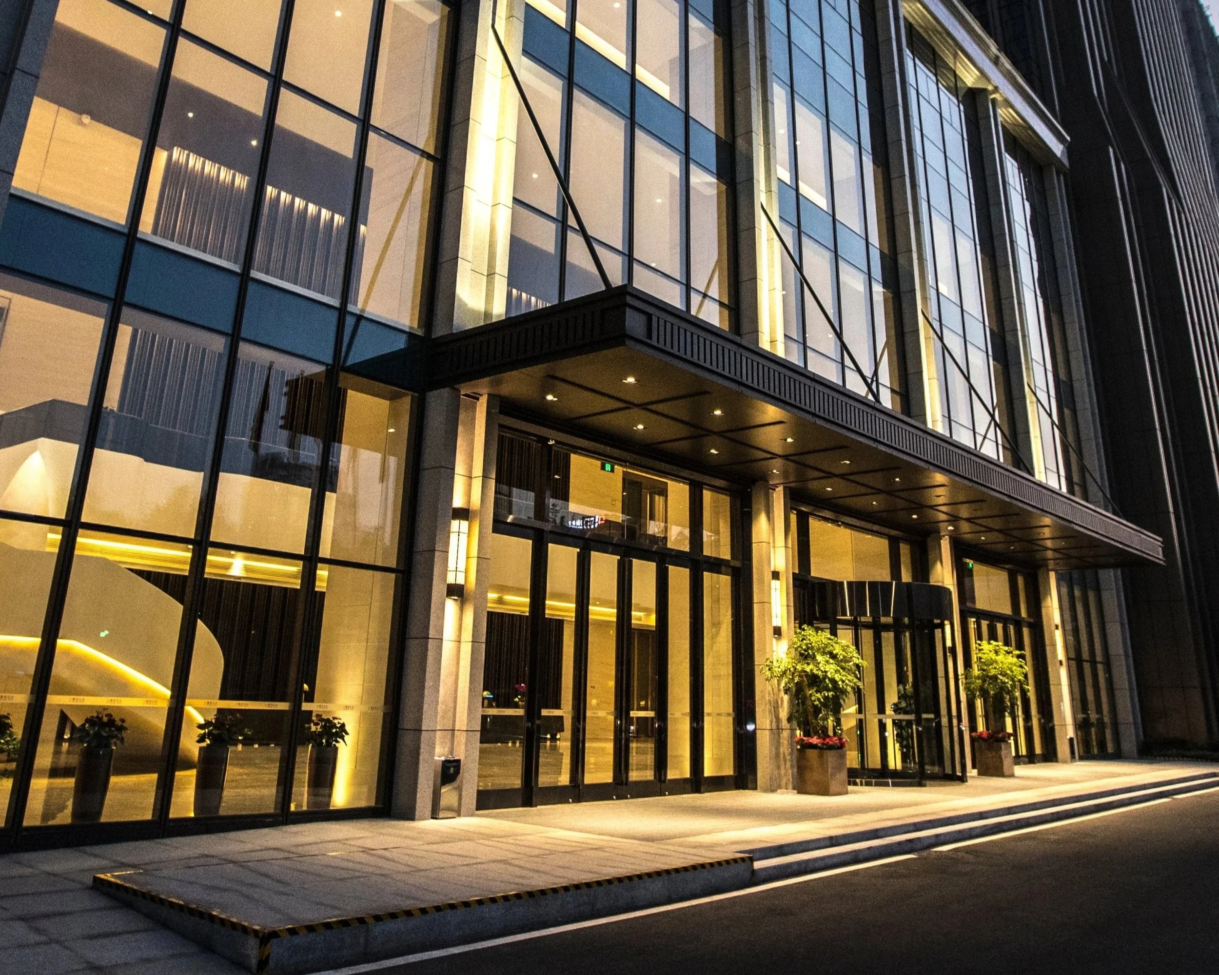 A modern multi-story building with a glass facade illuminated at night, featuring a covered entrance with outdoor lighting, planters, and trees at the front.