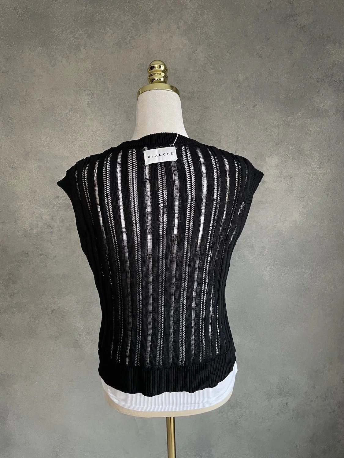 Back view of the black open-knit Blanche vest showing the vertical striped texture and white brand label at the inner collar.