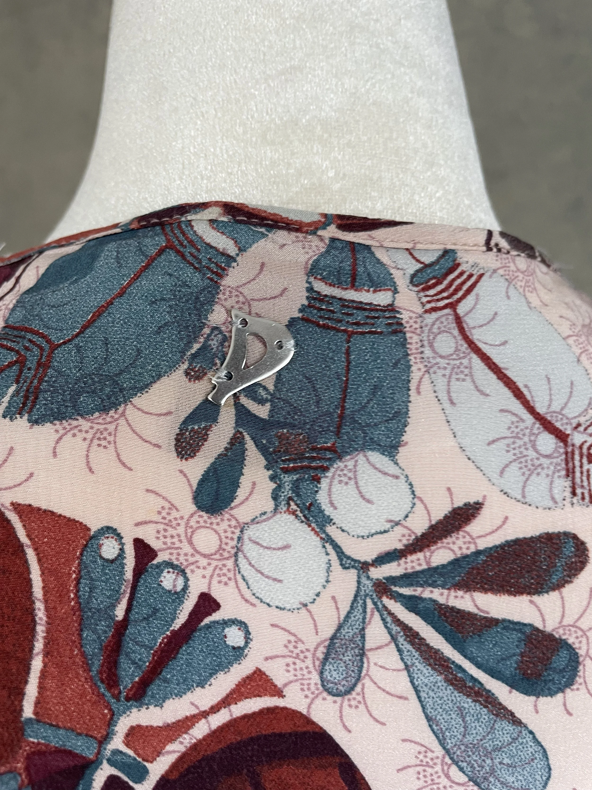 Macro detail of the small silver metal 'D' initial logo charm sewn onto the neck of the Dondup silk blouse.