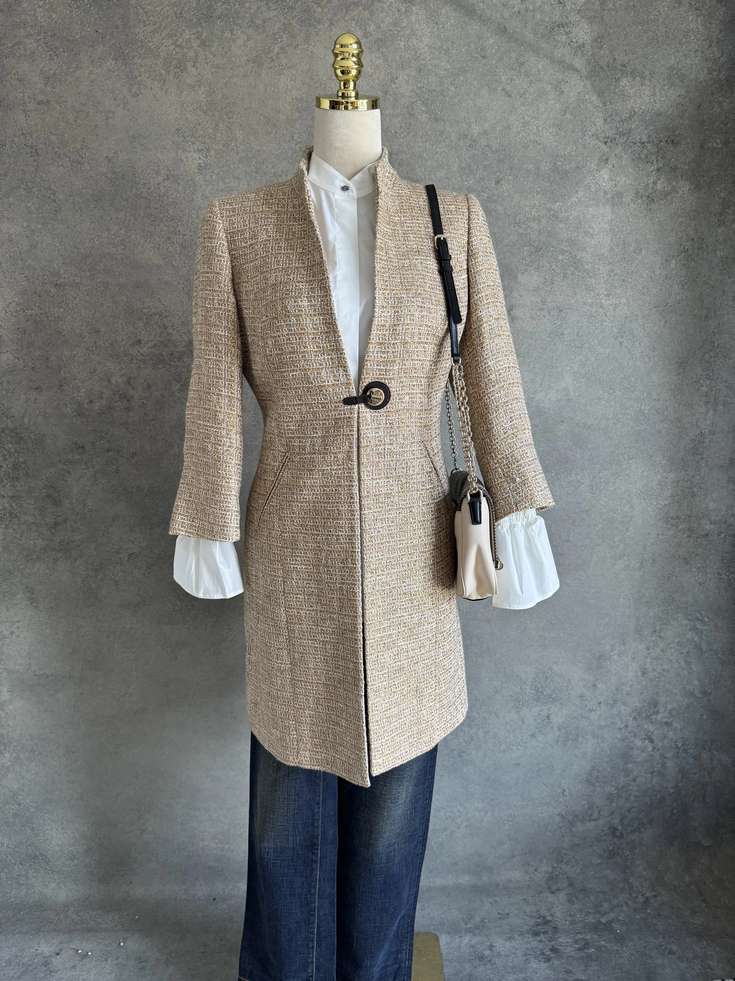 A mannequin dressed in a beige tweed coat over a white blouse with puffy sleeves, paired with dark jeans, and carrying a small beige purse with a chain strap against a gray textured background.