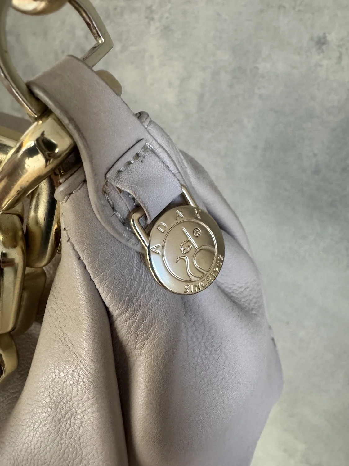 Close-up shot of gold Adax Since 1982 logo coin charm stitched onto soft beige leather