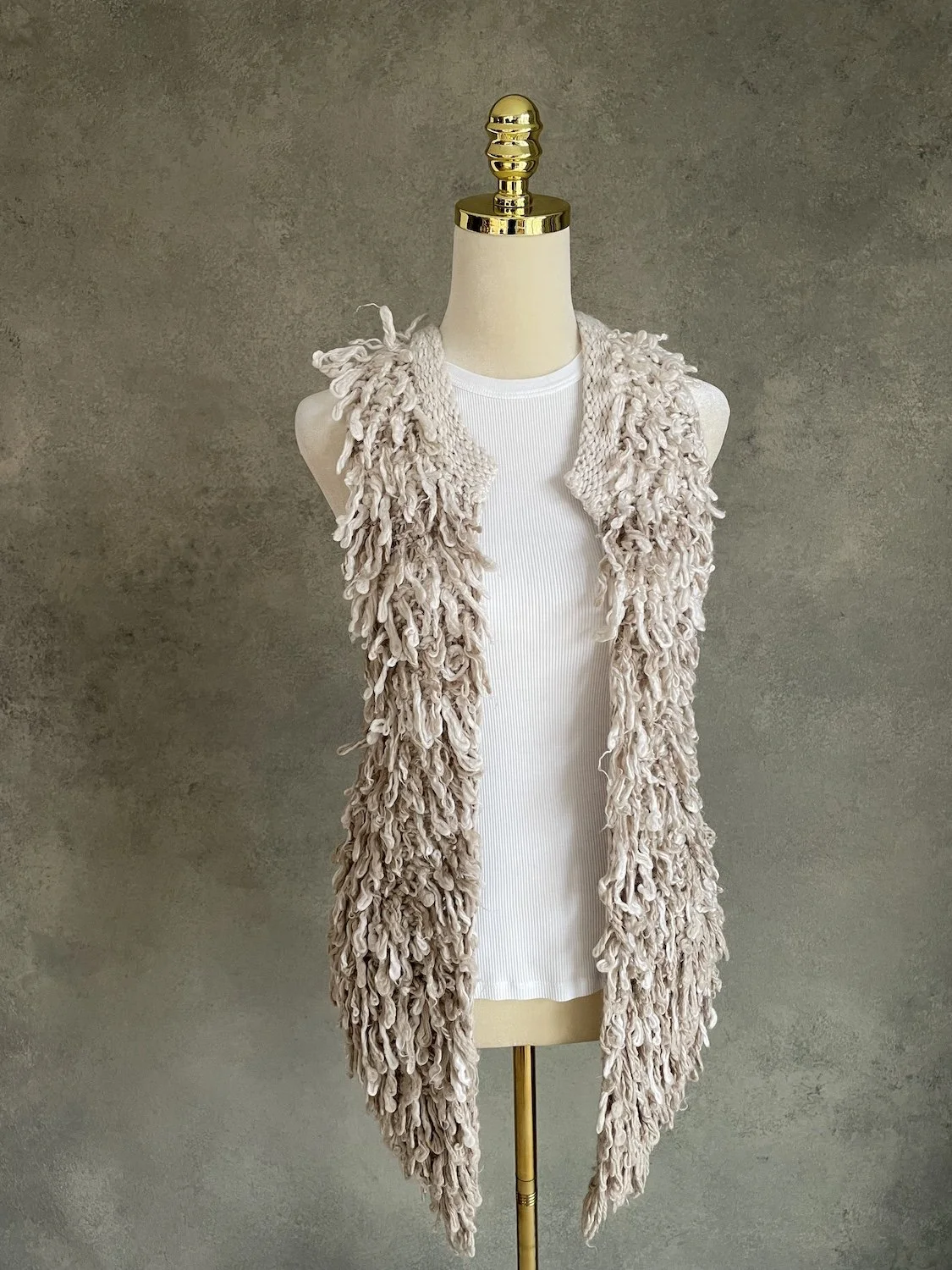 Front view of the greige shaggy knit vest on a mannequin against a grey concrete texture
