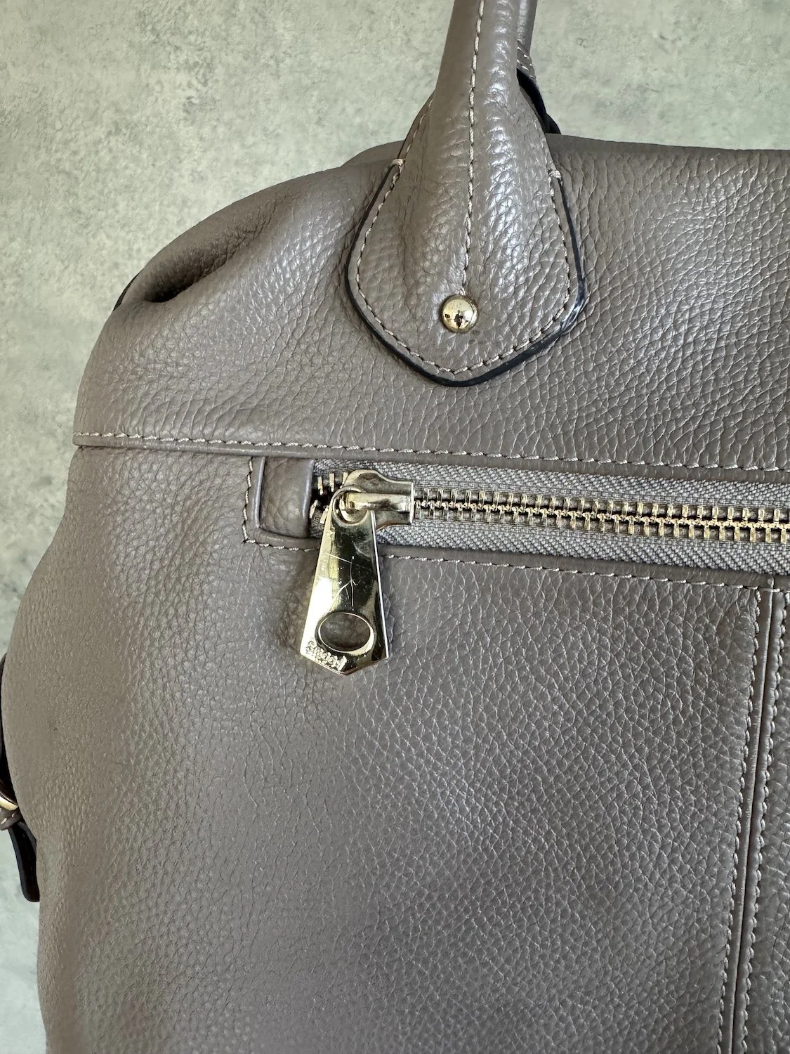 Close-up detail of gold-tone diamond Smaak logo tag attached to greige leather handle