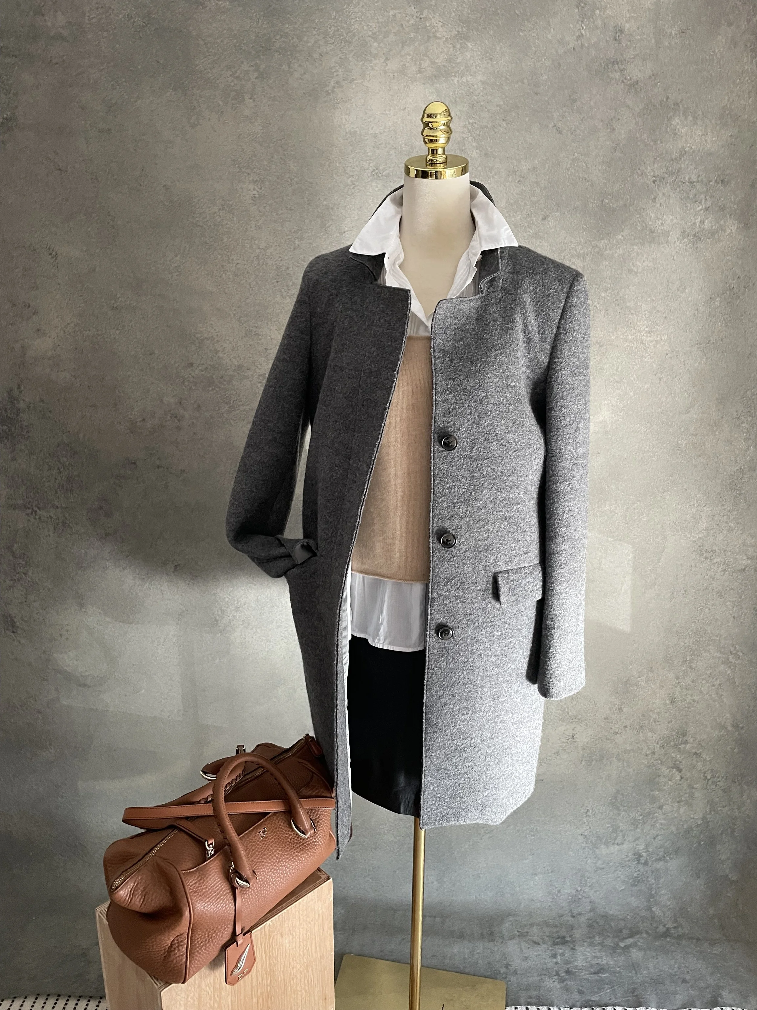 A mannequin dressed in a gray coat over a white shirt and beige sweater, upright against a neutral gray textured wall, with a brown handbag placed on a wooden stand in front.