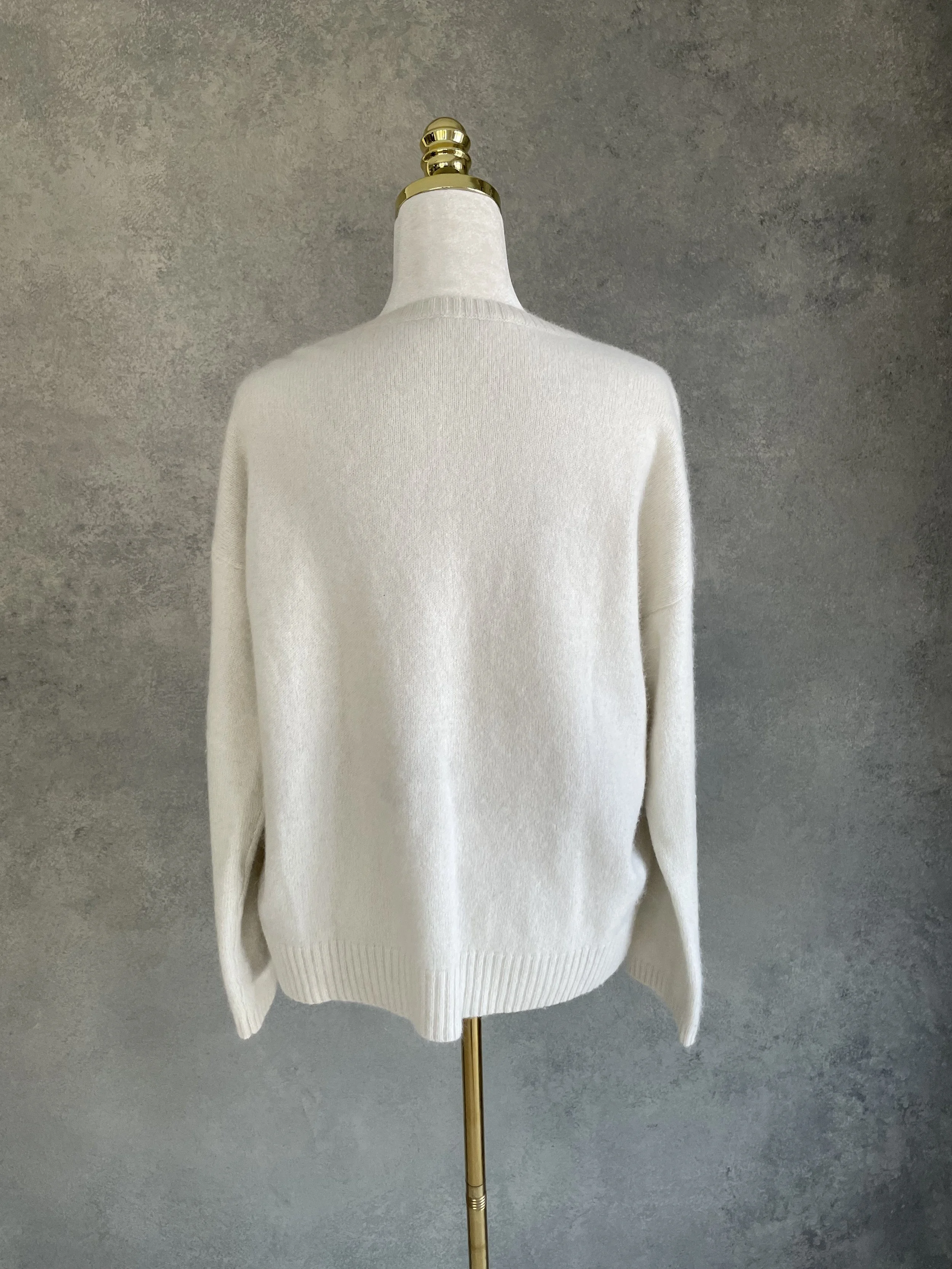 Back view of a Rue Blanche cream-colored round neck sweater on a mannequin against a grey textured background.