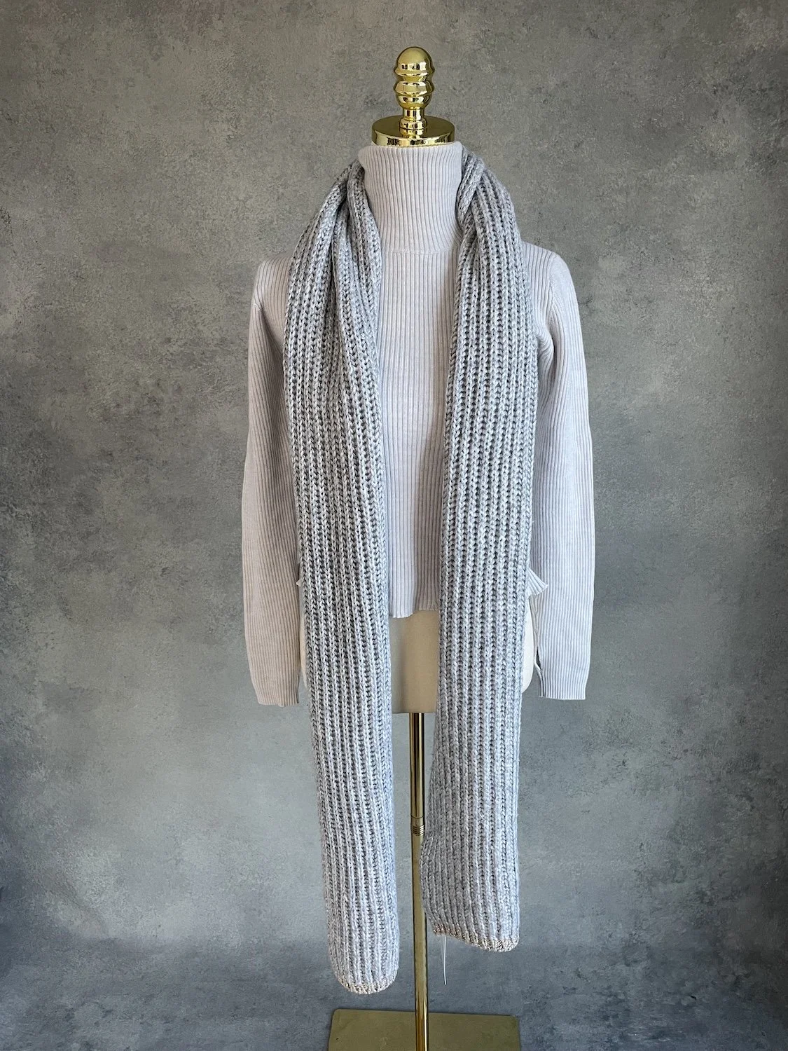 Xandres grey ribbed knit scarf with gold trim draped on a mannequin over a light turtleneck against a neutral textured background.