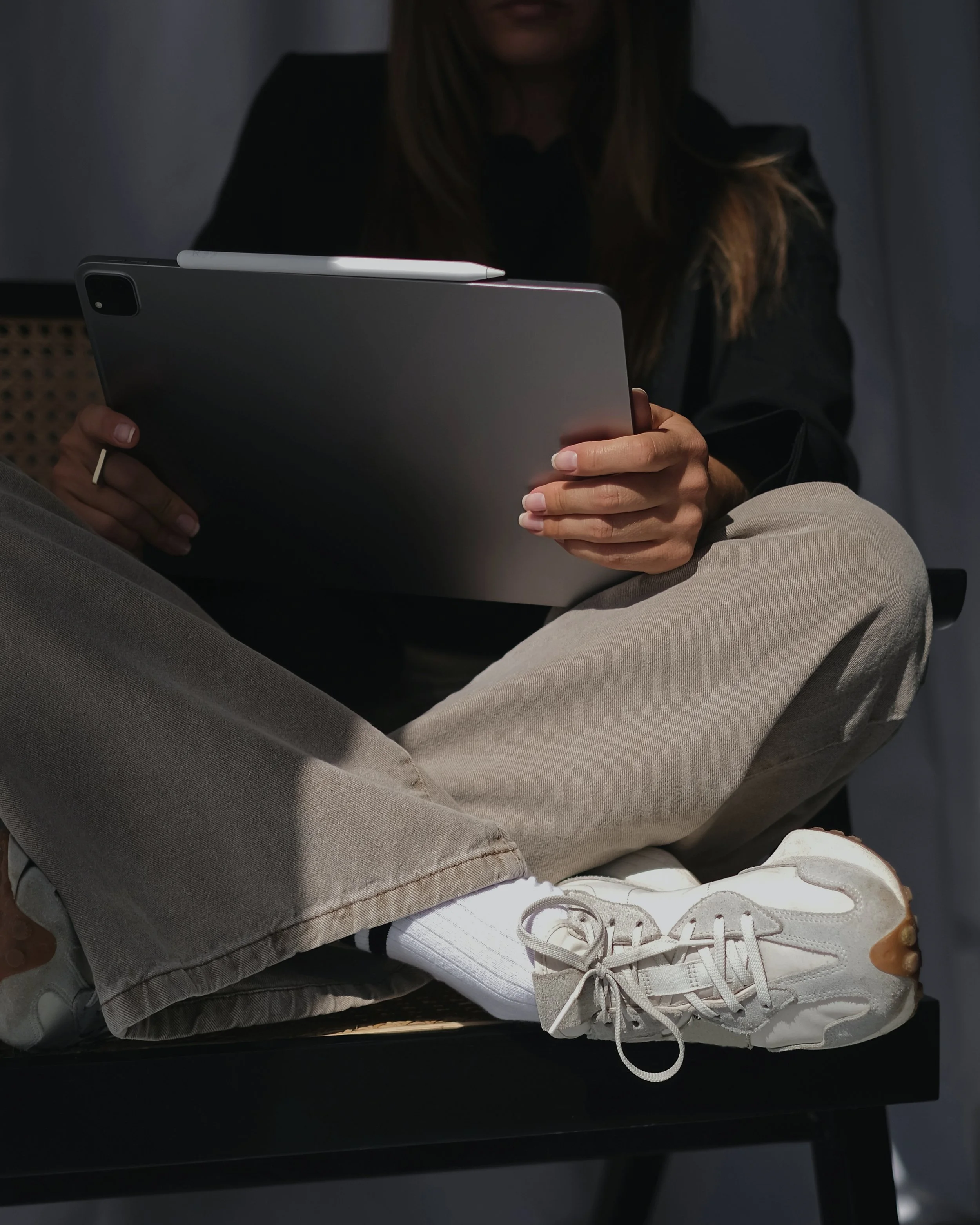 Person sitting with crossed legs wearing beige sneakers, beige pants, holding a tablet with a white stylus resting on it, in a dimly lit room.