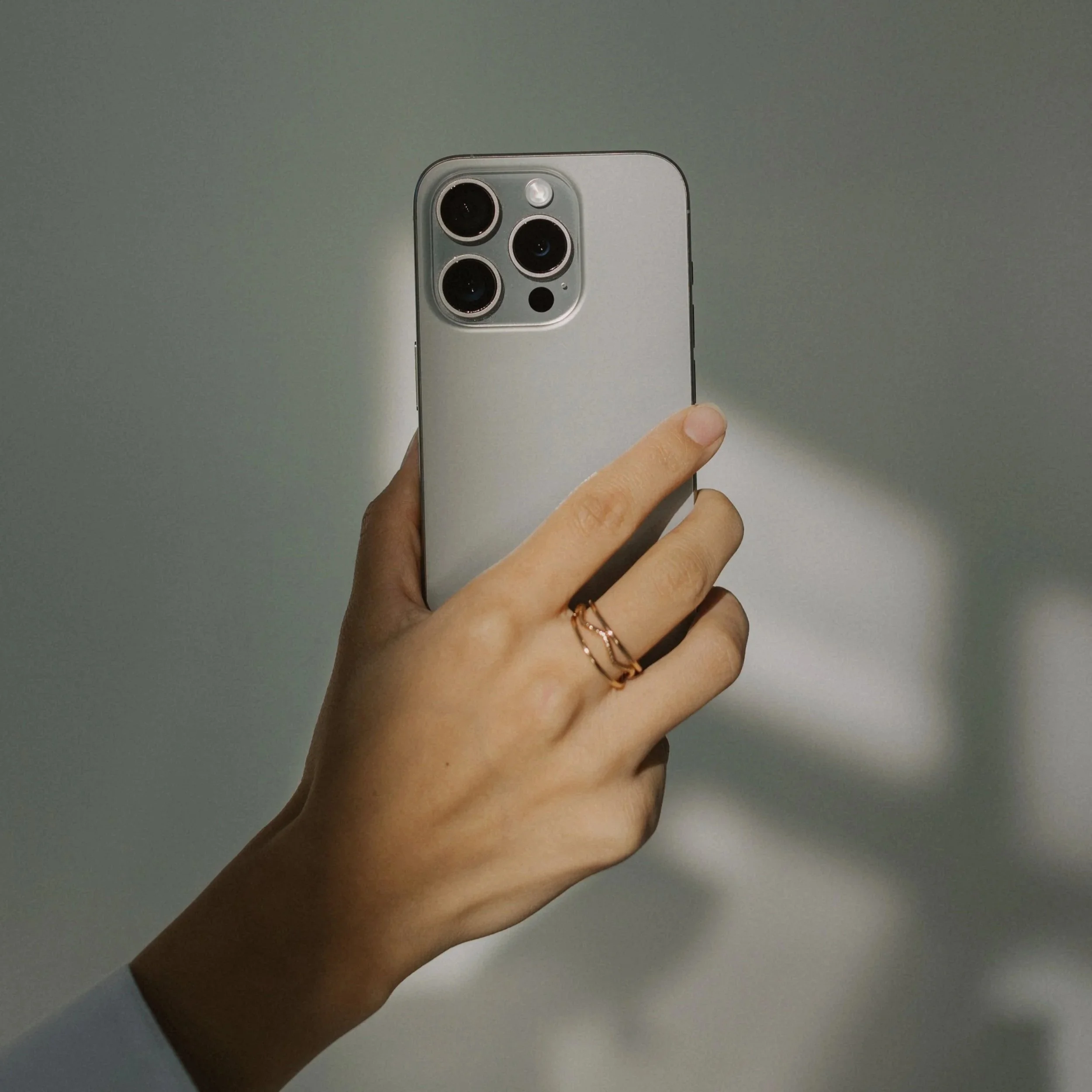 Close-up of a person's hand holding a silver smartphone with three camera lenses on the back, against a plain grey background.