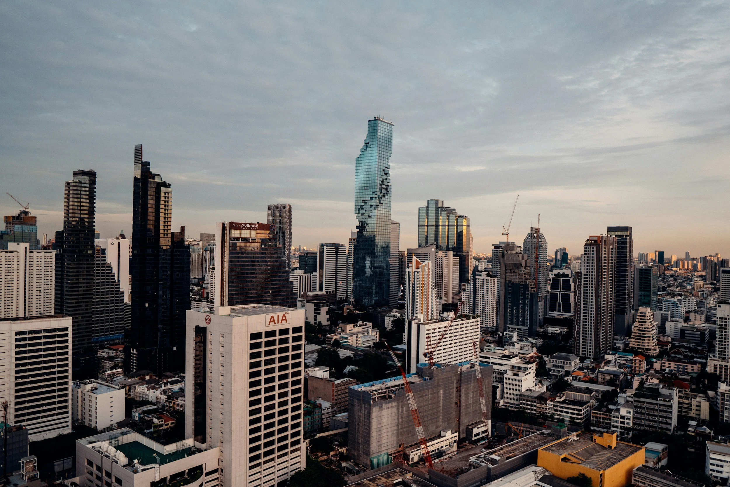 Skyline of a city with tall buildings, some under construction, and a uniquely shaped modern glass skyscraper with a spiral design, under a partly cloudy sky.