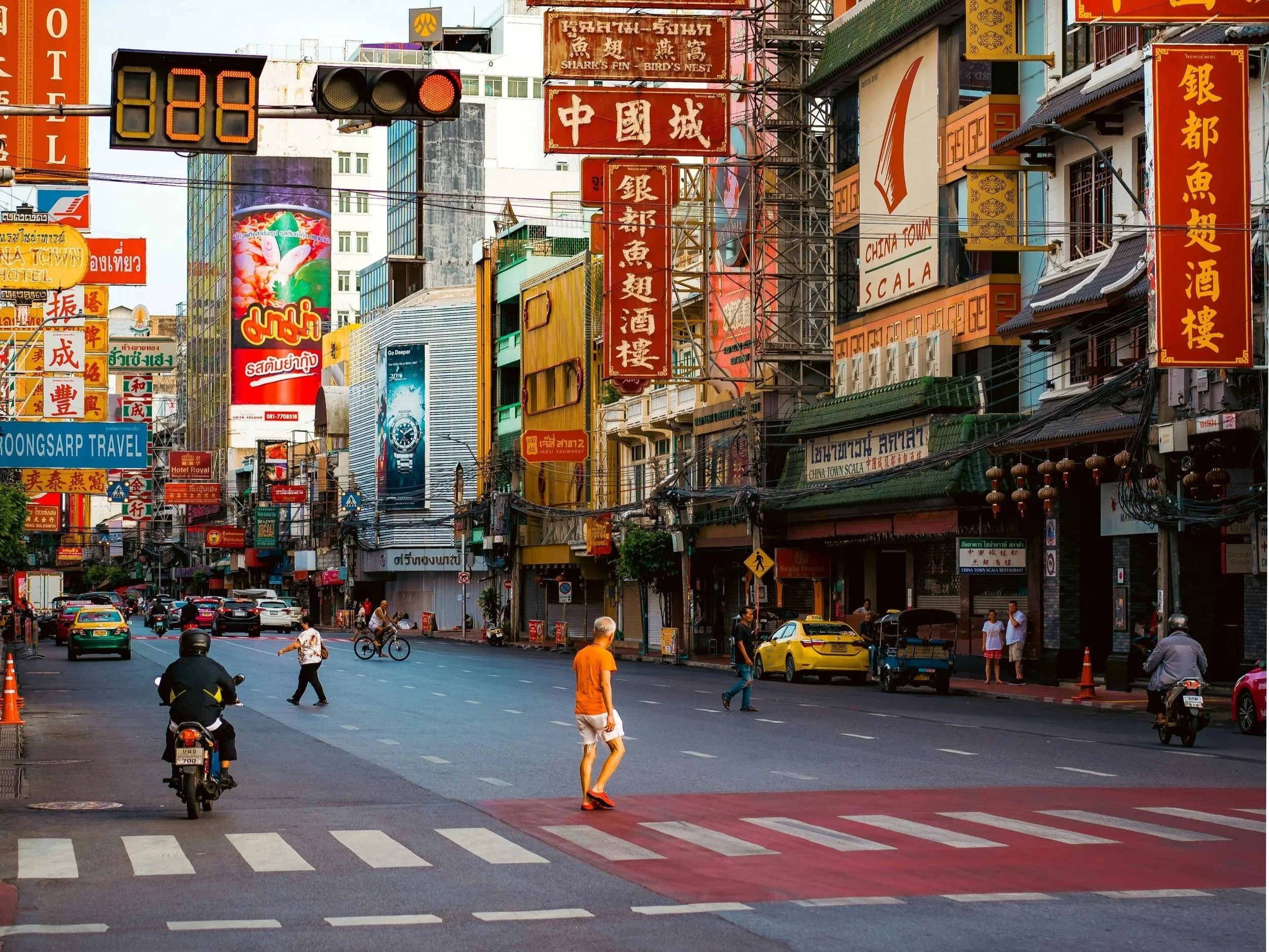Street scene in Chinatown with many colorful signs in Chinese and Thai, pedestrians crossing the street, and cars parked along the sides. There are several buildings with neon signs and a traffic light showing closed at the intersection.