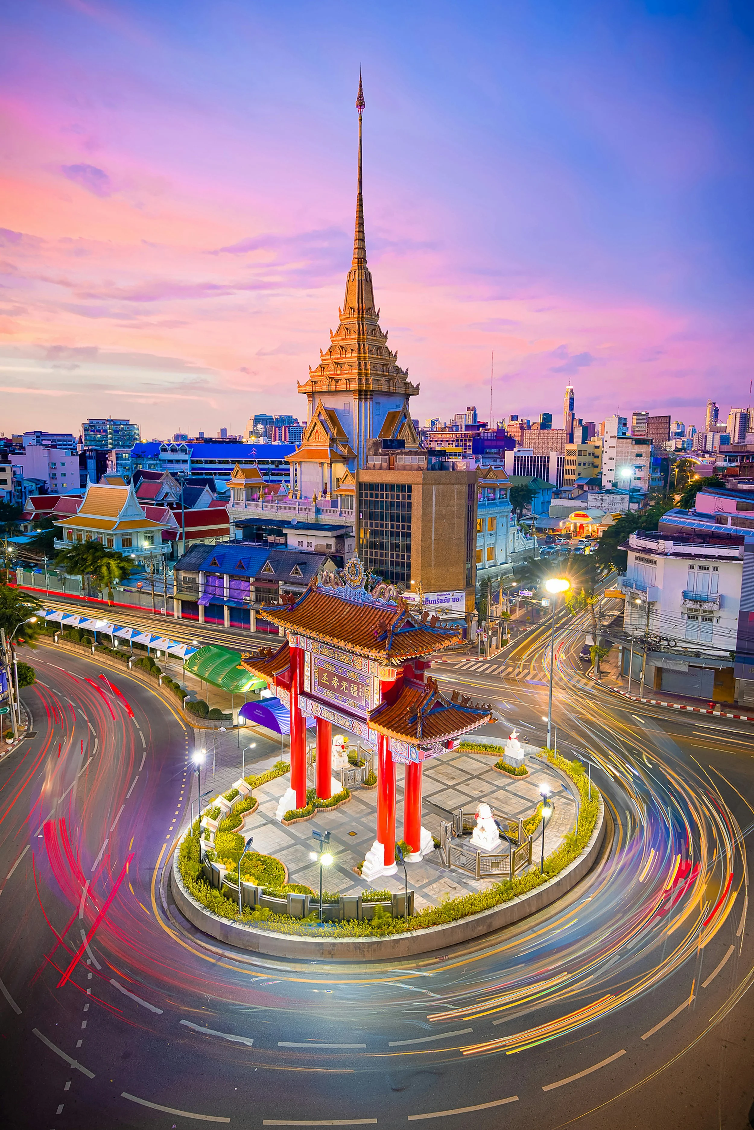 A cityscape at sunset with a prominent traditional Asian-style tower and a roundabout with colorful light trails from moving vehicles.