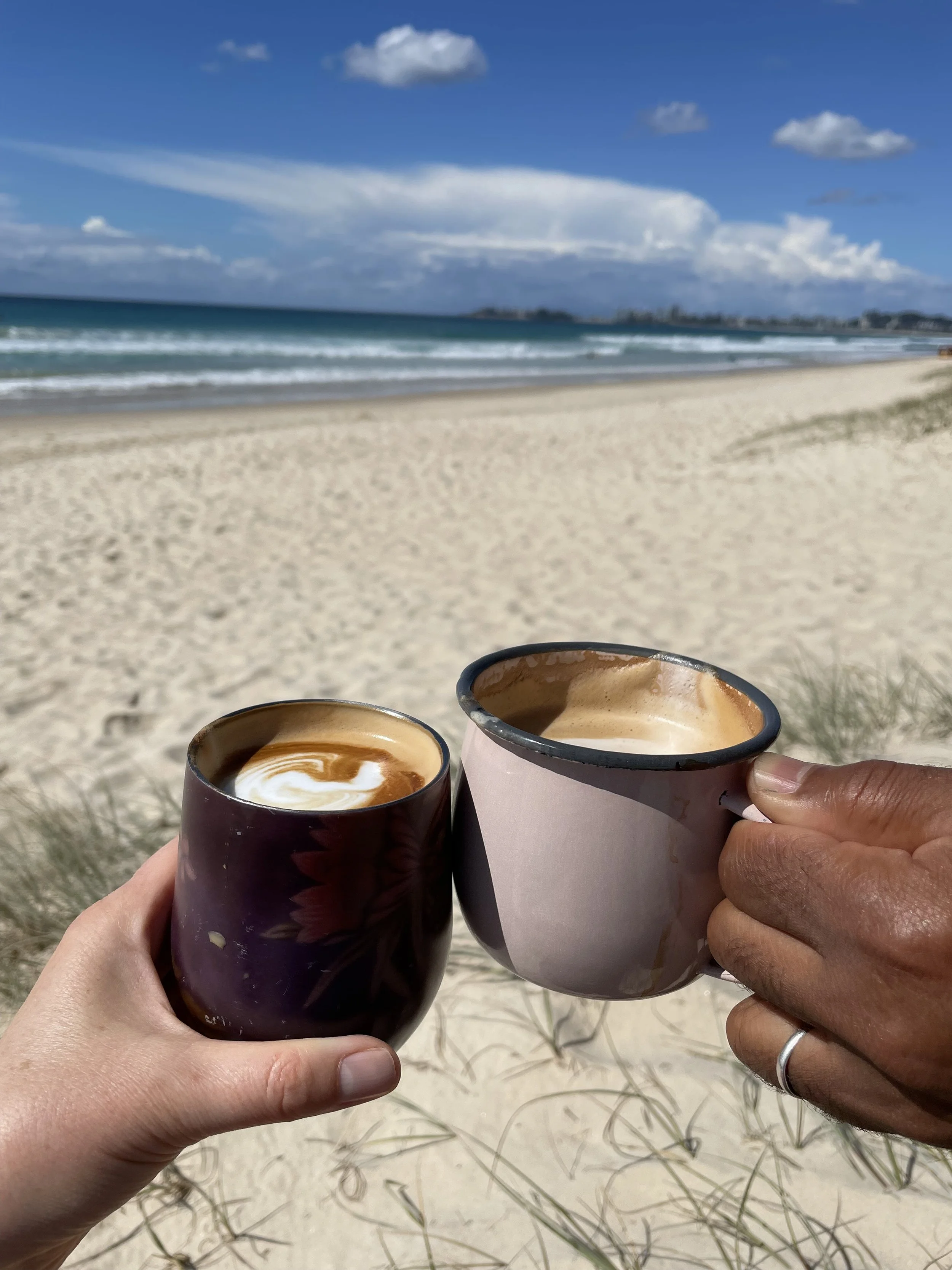 2 people holding coffee in cups, beach view.