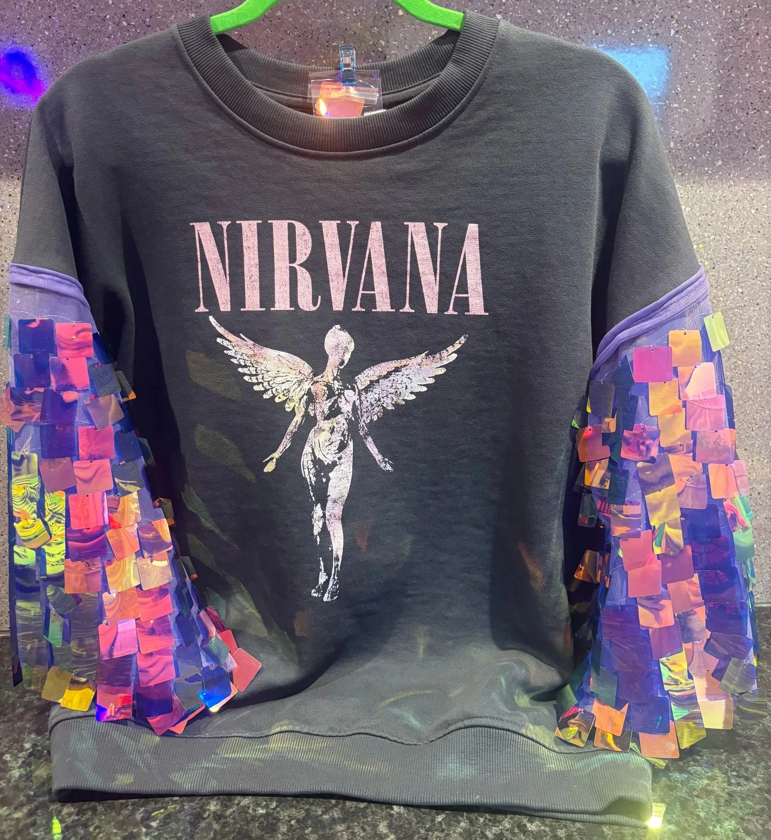 Black Nirvana band sweatshirt with a graphic of a moth and pink band logo, displayed on a green hanger with iridescent square roll-up sleeves, on a dark speckled surface.