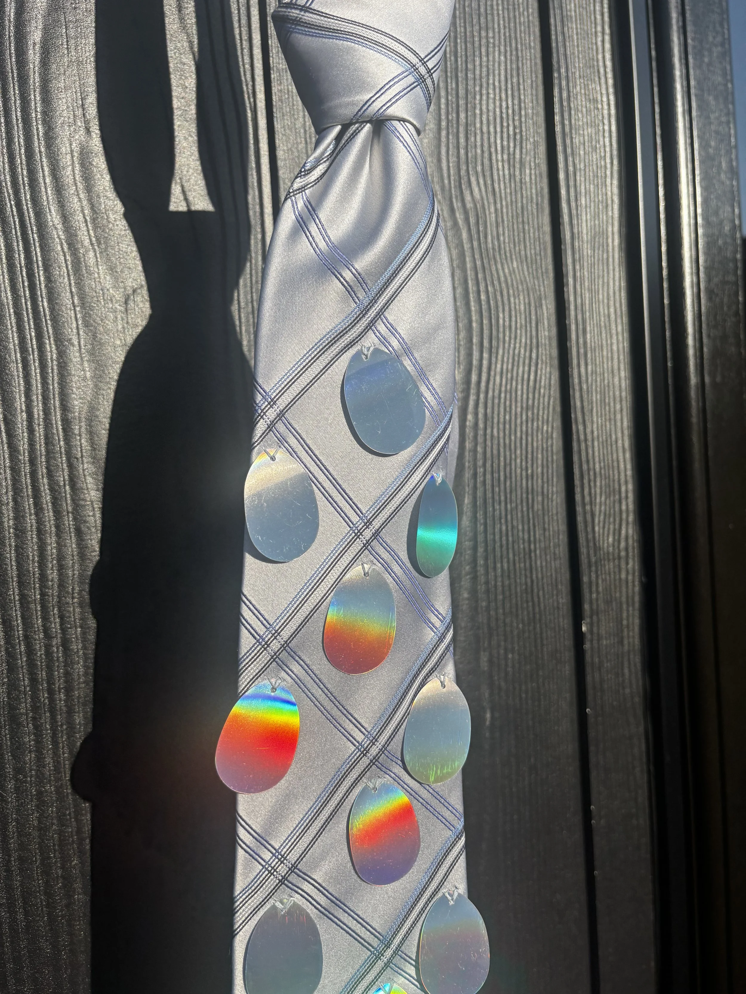 Close-up of a strip of reflective, holographic stickers with rainbow colors, partially covered by a gray silk tie with blue lines, placed on a black textured surface.
