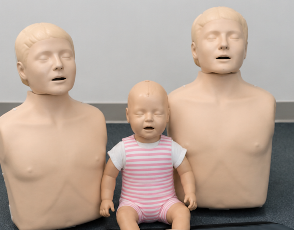 A baby doll and two mannequin torsos with closed eyes and open mouths, placed on a dark surface against a plain light-colored wall.