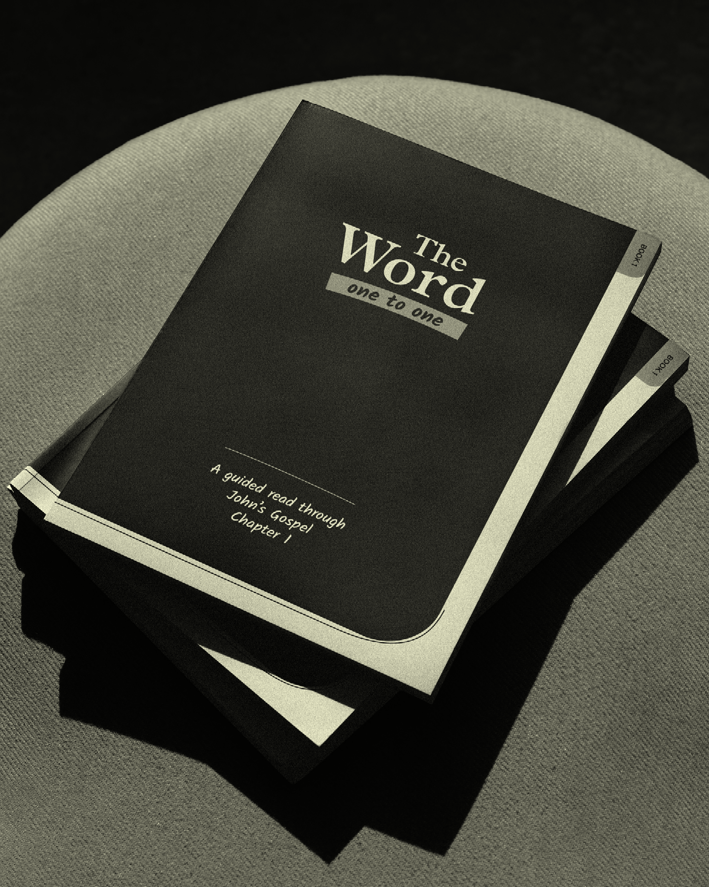 A black booklet titled 'The Word' with subtitle 'one to one' on a light gray fabric surface. Small tabs indicate sections labeled 'book 1' and 'book 2'.