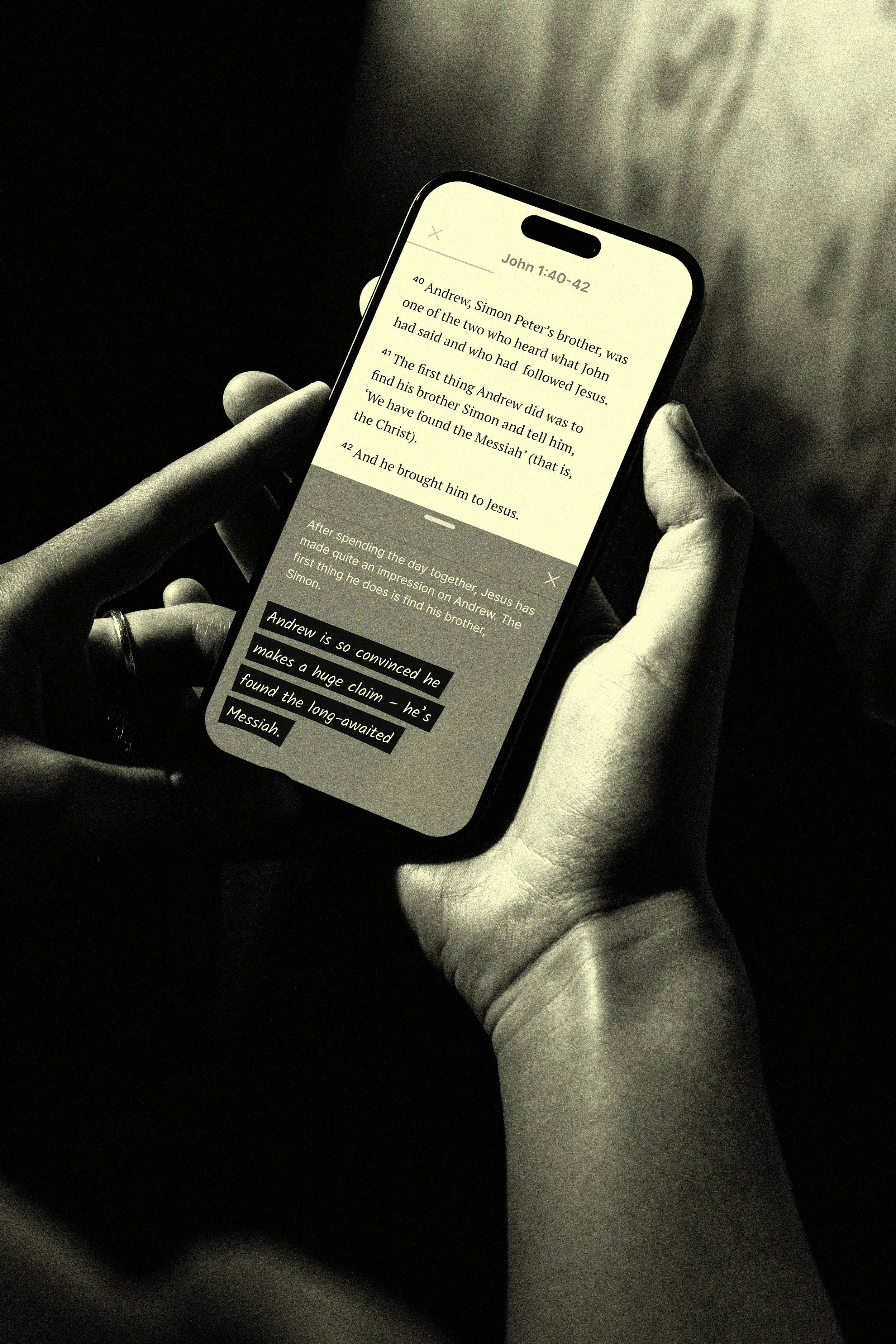 A person holding a smartphone displaying a Bible passage from John 1:40-42, with black and white tones and a dark background.