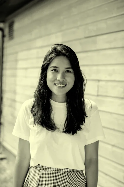 Black and white photo of a young woman with long hair smiling, standing in front of a wooden wall.