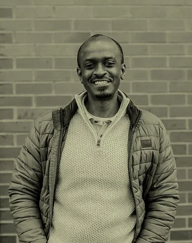 A smiling man standing outdoors in front of a brick wall, dressed in a quilted jacket and hoodie.